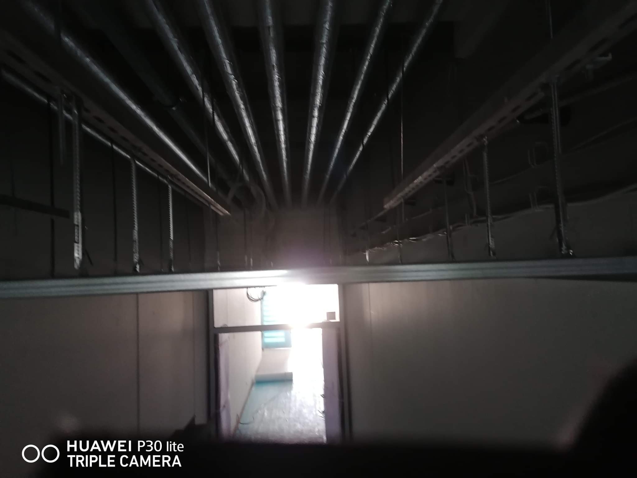 Metalowe rury pod sufitem w pomieszczeniu, widok na jasne pomieszczenie przez drzwi, instalacja elektryczna w trakcie montażu.