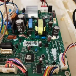 Zielona płyta główna urządzenia AGD z widocznymi kondensatorami, transformatorami i złączami kablowymi, umieszczona w obudowie z tworzywa sztucznego.