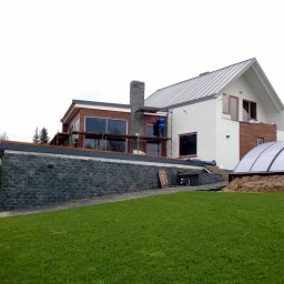 Zakład Budowlany Zygmunt Formela - Dom jednorodzinny w trakcie prac wykończeniowych elewacji, widoczne rusztowania i pracownik w niebieskim ubraniu przy kominie, zadaszenie basenu w tle.