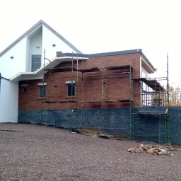 Zakład Budowlany Zygmunt Formela - Dom w trakcie wykończenia elewacji: fragment ściany z ciemnego kamienia, wyżej drewniana elewacja w rusztowaniach, widoczne okna i schody zewnętrzne.