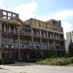 Zakład Budowlany Zygmunt Formela - Budynek w trakcie wznoszenia z betonu komórkowego, widoczne drewniane rusztowania i podpory, na tle błękitnego nieba i innego budynku mieszkalnego.