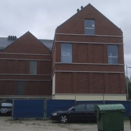 Zakład Budowlany Zygmunt Formela - Nowoczesny budynek z czerwonej cegły z szarymi elementami dekoracyjnymi, widoczny z poziomu ziemi, z zaparkowanymi samochodami i pojemnikami na śmieci z przodu.