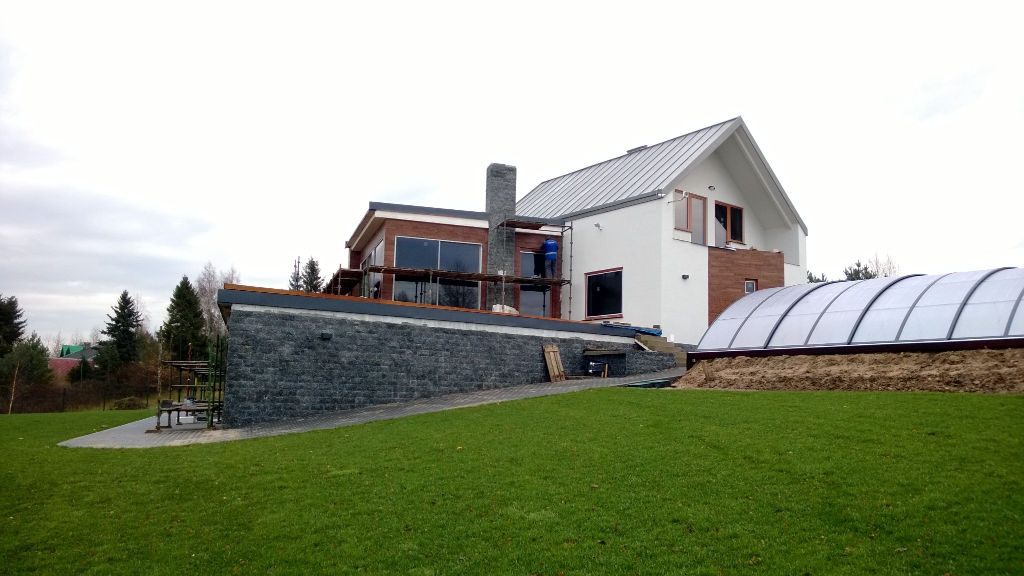 Dom jednorodzinny w trakcie prac wykończeniowych elewacji, widoczne rusztowania i pracownik w niebieskim ubraniu przy kominie, zadaszenie basenu w tle.