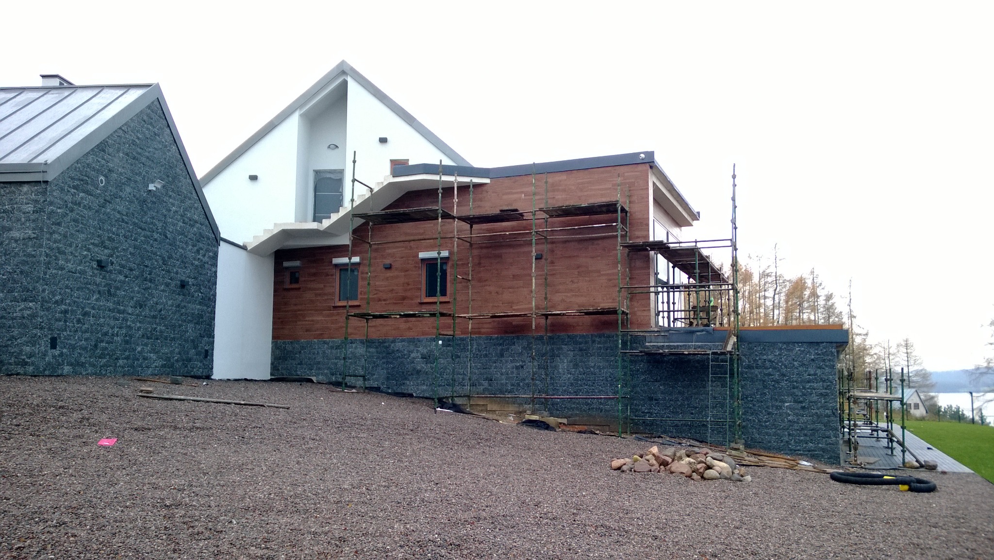 Dom w trakcie wykończenia elewacji: fragment ściany z ciemnego kamienia, wyżej drewniana elewacja w rusztowaniach, widoczne okna i schody zewnętrzne.