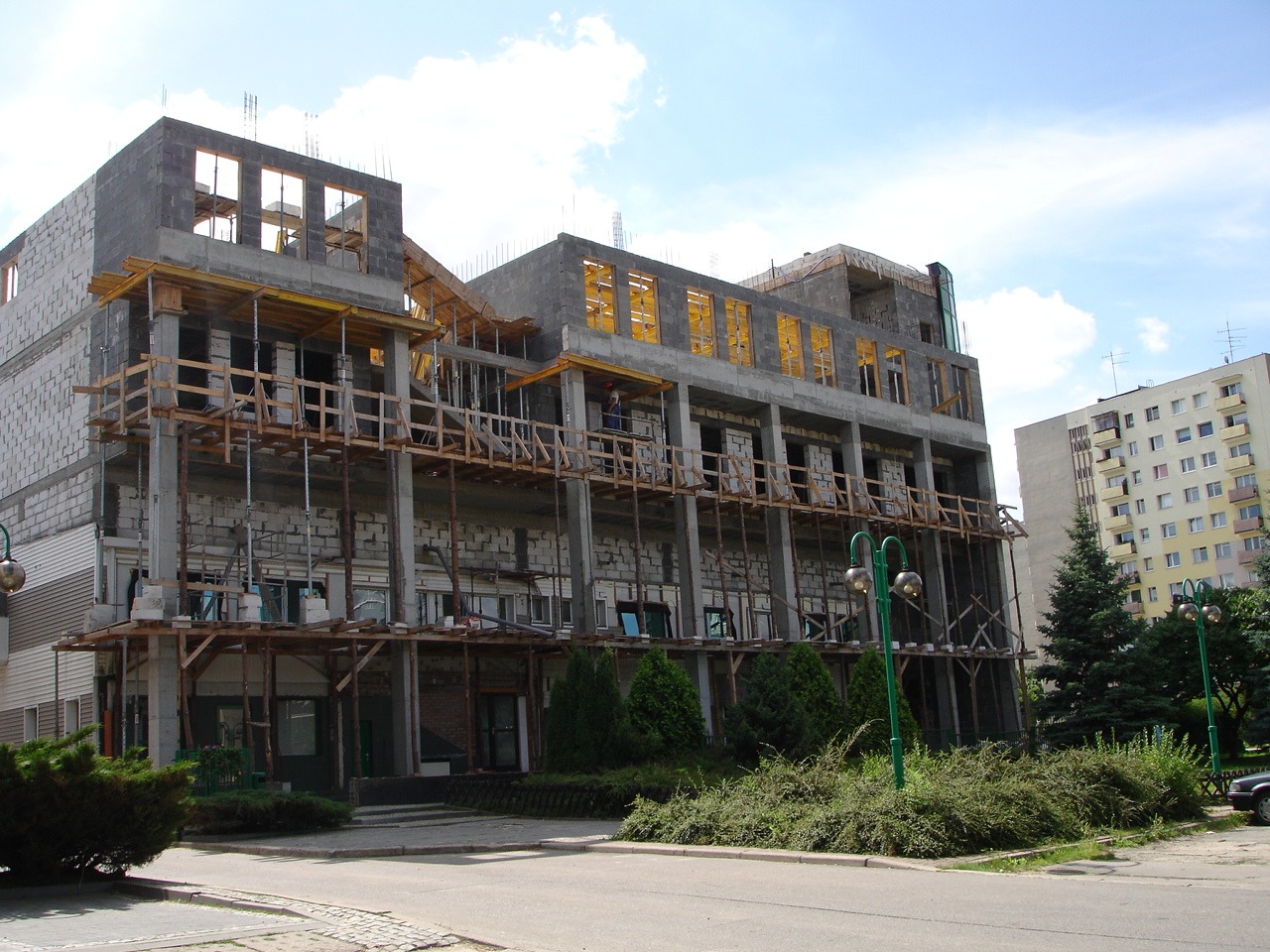 Budynek w trakcie wznoszenia z betonu komórkowego, widoczne drewniane rusztowania i podpory, na tle błękitnego nieba i innego budynku mieszkalnego.