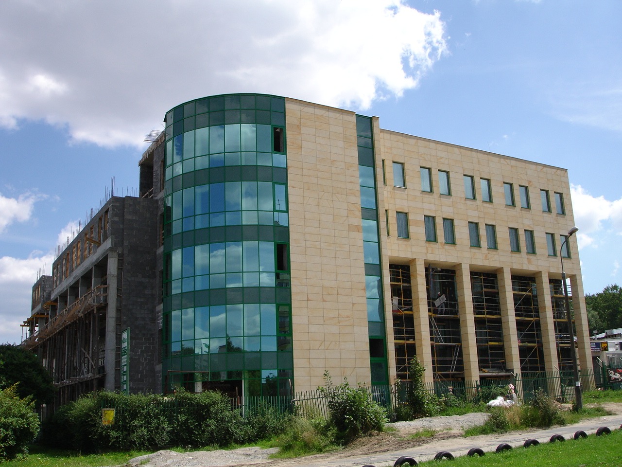 Budynek biurowy w trakcie budowy z widoczną konstrukcją żelbetową, fragmentem elewacji z piaskowca i przeszkloną, zaokrągloną fasadą.