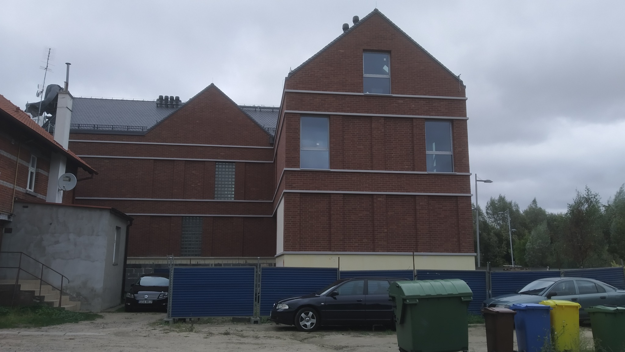 Nowoczesny budynek z czerwonej cegły z szarymi elementami dekoracyjnymi, widoczny z poziomu ziemi, z zaparkowanymi samochodami i pojemnikami na śmieci z przodu.