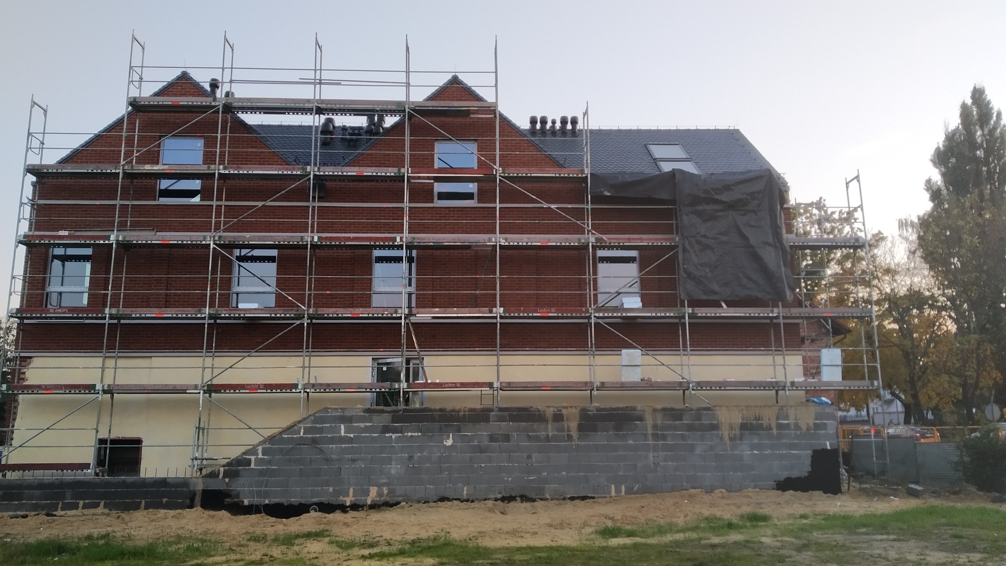 Wielopiętrowy budynek z czerwonej cegły w trakcie prac elewacyjnych, otoczony rusztowaniami, z widoczną częścią ocieploną i fragmentem murowanej ściany z bloczków betonowych w dolnej partii.