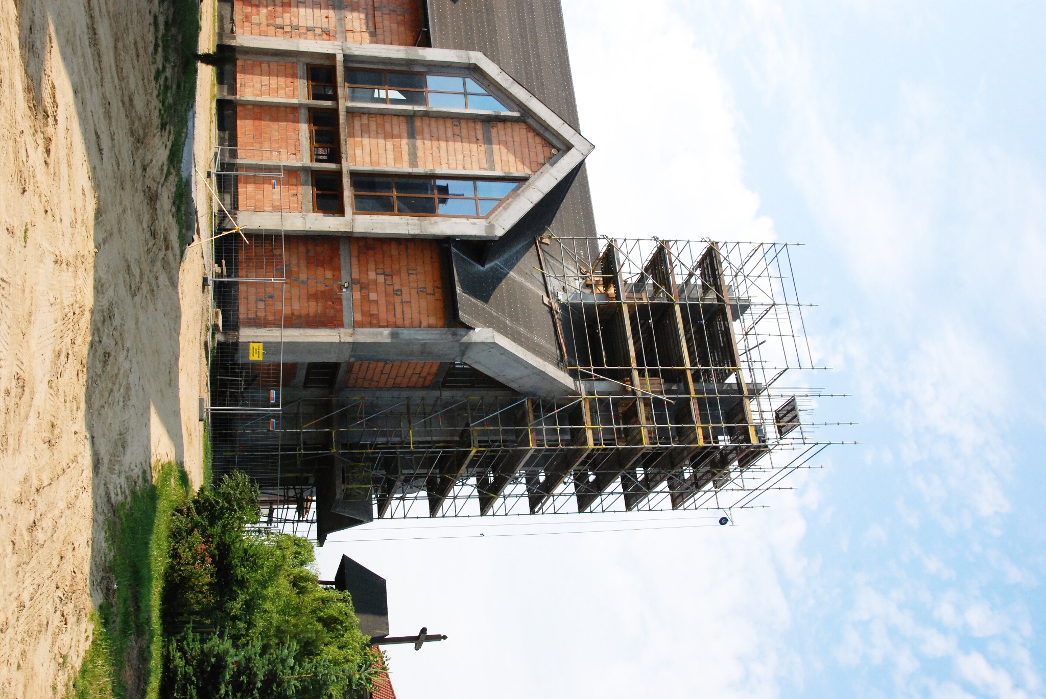 Budynek w trakcie budowy z cegły i betonu, otoczony rusztowaniami, widok z dołu na tle jasnego nieba.