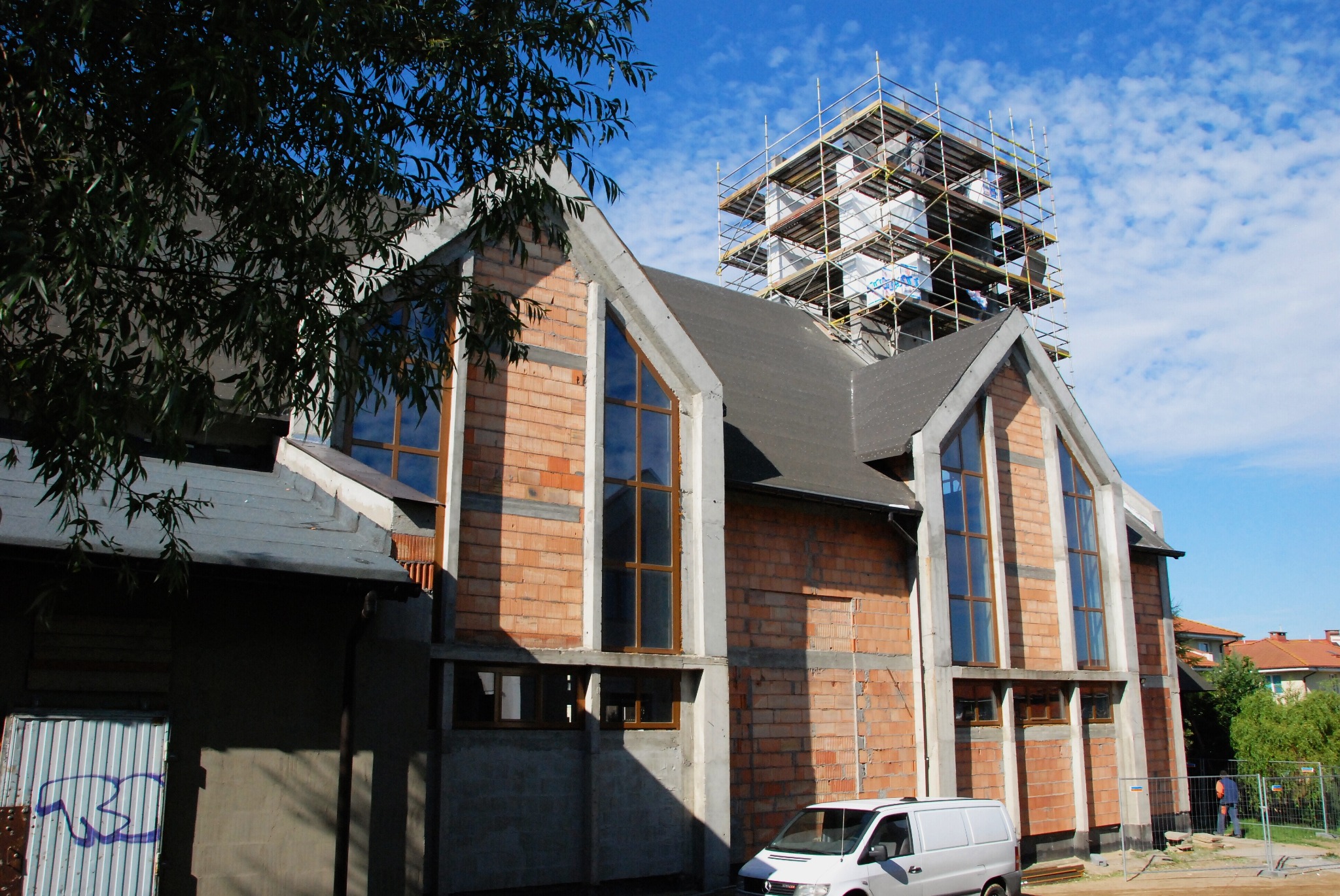 Budynek w trakcie murowania ścian z czerwonej cegły, z wysokimi oknami i szarymi elementami konstrukcyjnymi, otoczony rusztowaniem, z widocznym białym vanem i fragmentem drzewa w kadrze.