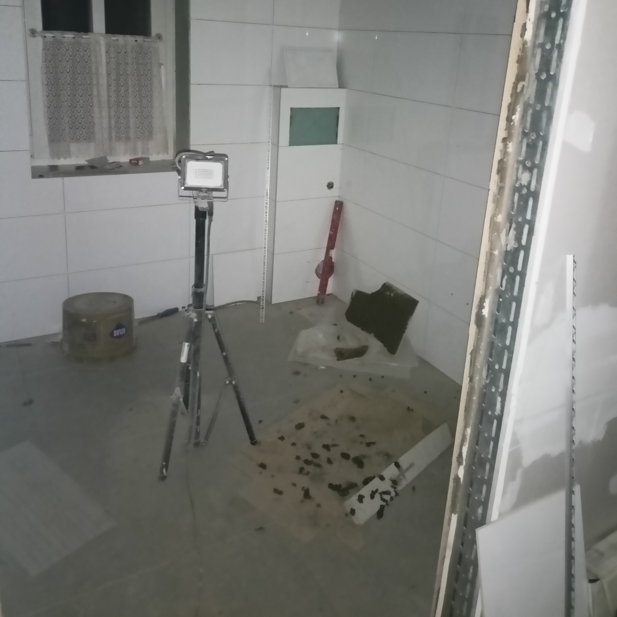 Wnętrze łazienki w trakcie remontu: białe płytki na ścianach, widoczna puszka Atlas, poziomica, reflektor na statywie, płyta kartonowo-gipsowa na metalowym stelażu.