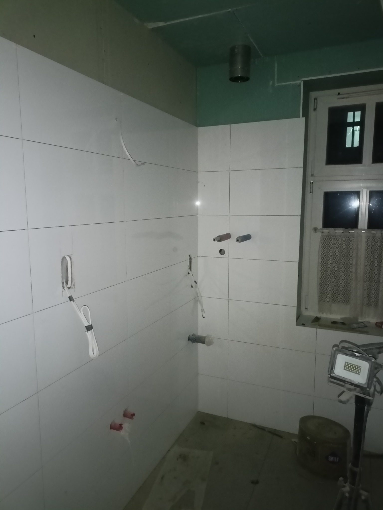 Łazienka w trakcie remontu: białe płytki na ścianach, widoczne wystające rury, kable elektryczne, surowy sufit z płyt karton-gips i lampa budowlana na statywie.