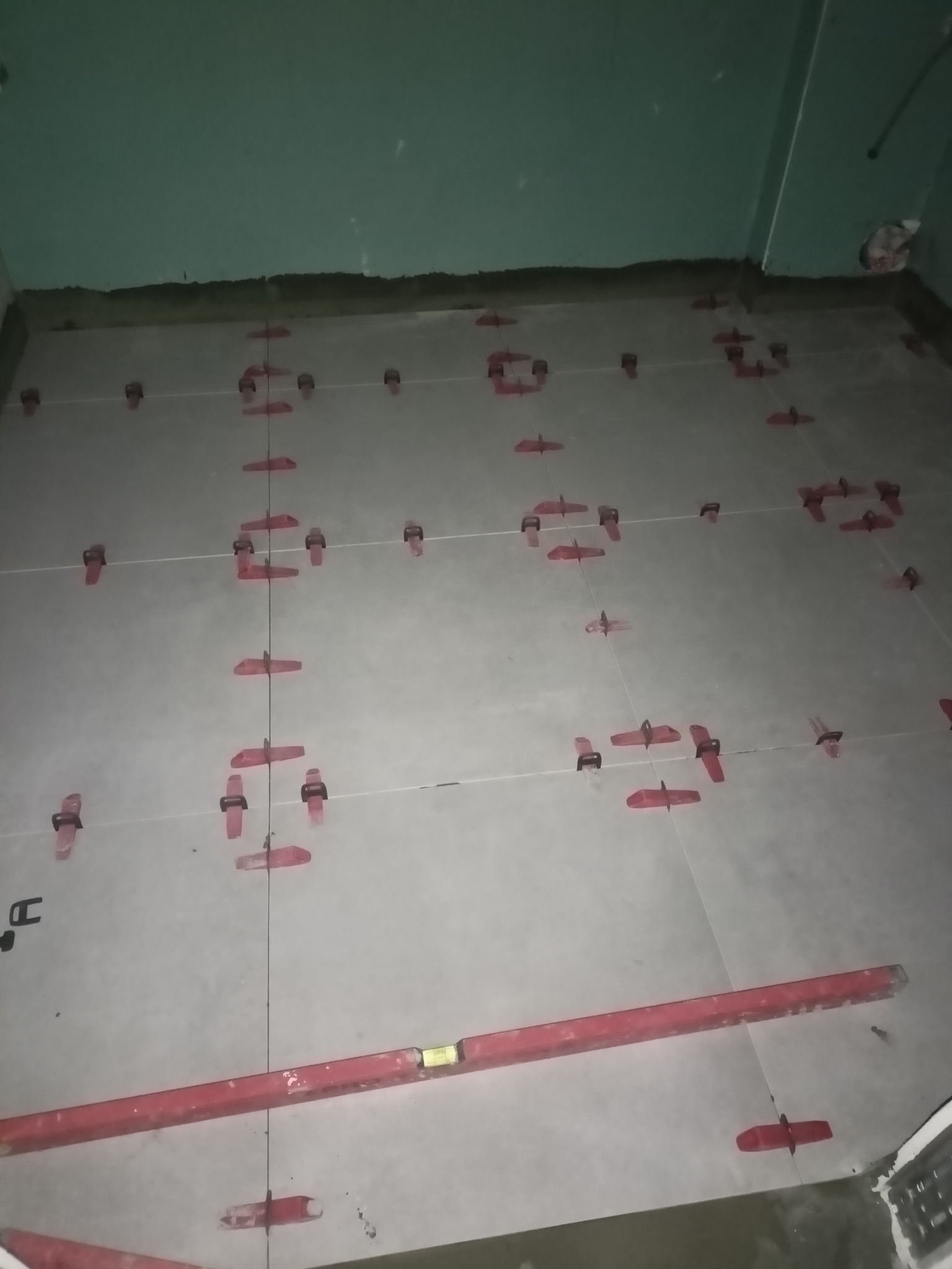 Podłoga w trakcie układania płytek, widoczne krzyżyki dystansowe i poziomica, surowy stan ścian