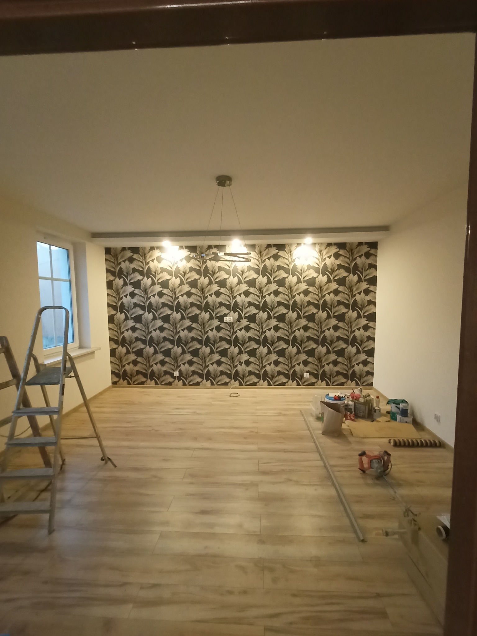Pokój po remoncie z drewnianą podłogą i ścianą pokrytą tapetą w liście, widoczna drabina i narzędzia malarskie.