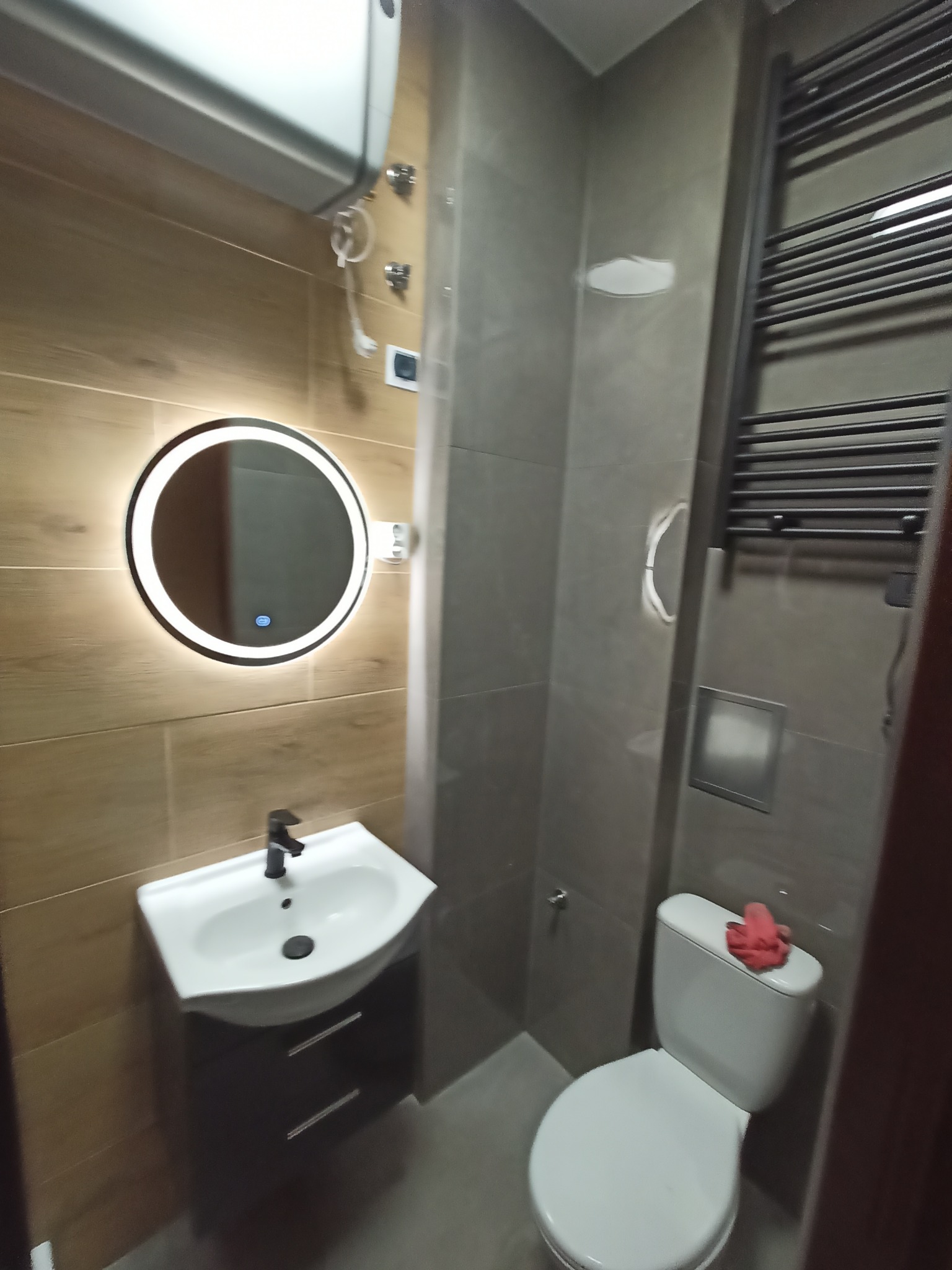 Nowoczesna łazienka z drewnopodobnymi płytkami, okrągłym lustrem z podświetleniem LED, wiszącą szafką z umywalką i grzejnikiem drabinkowym w kolorze grafitowym.