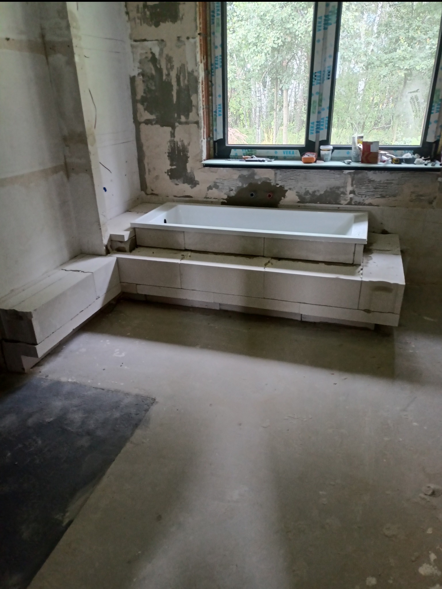 Wanna w trakcie obudowy z bloczków betonowych w surowej łazience, widoczne okno i przygotowane ściany.