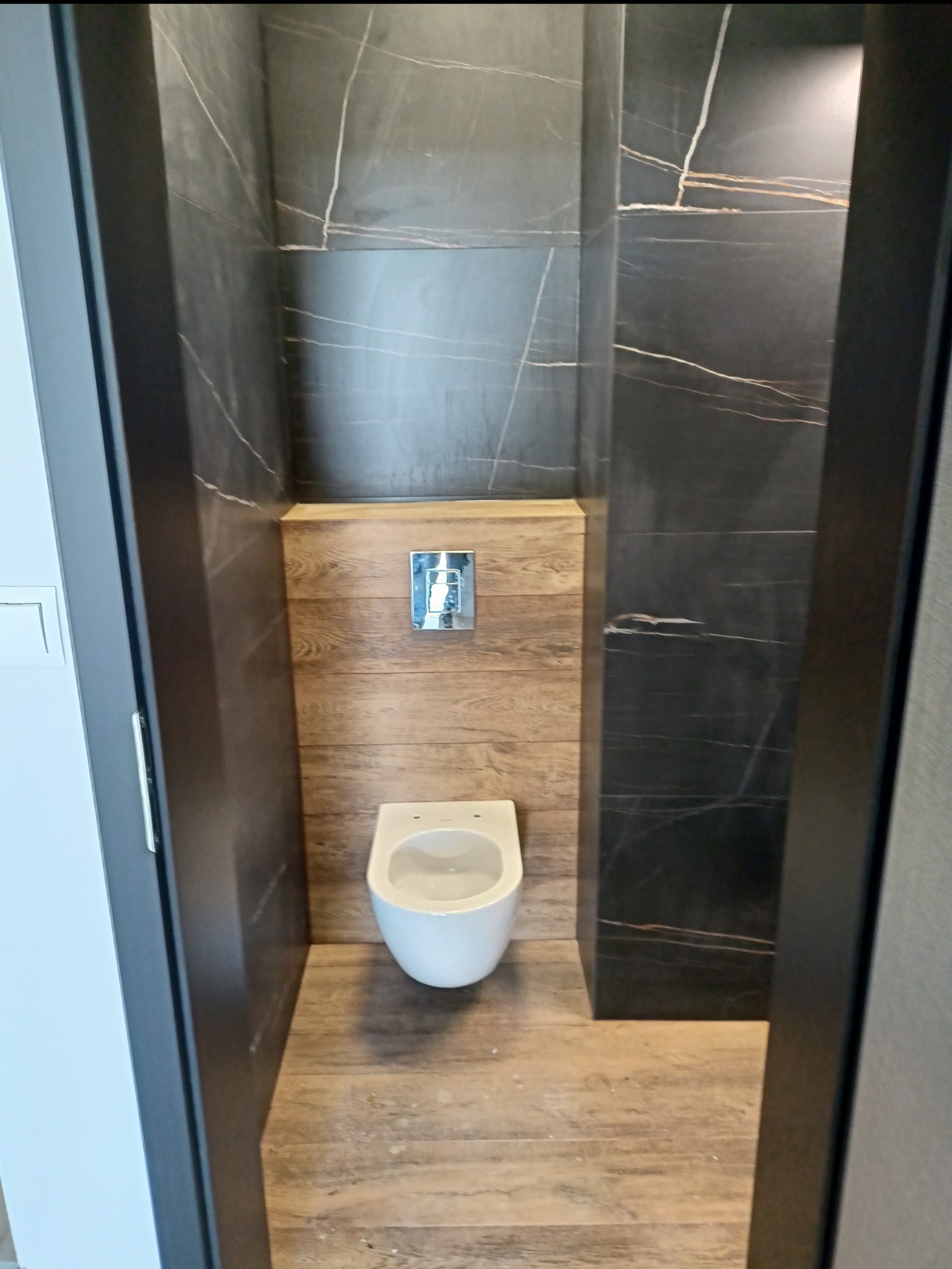 Nowoczesna toaleta podwieszana z drewnopodobną zabudową i czarnymi płytkami imitującymi marmur z jasnymi żyłami.