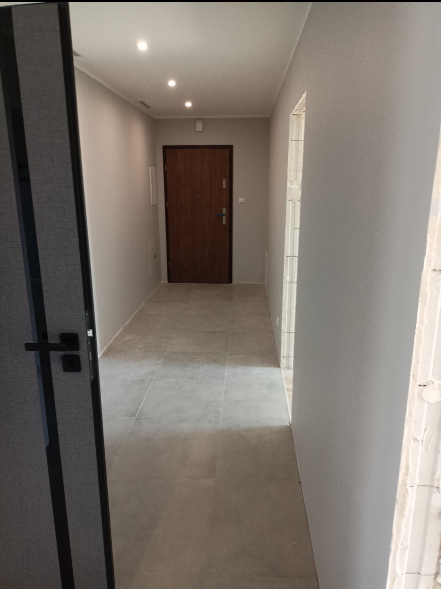 Korytarz po remoncie z szarymi ścianami, nową podłogą z płytek i widocznym ościeżem drzwi w trakcie montażu, światło punktowe na suficie.