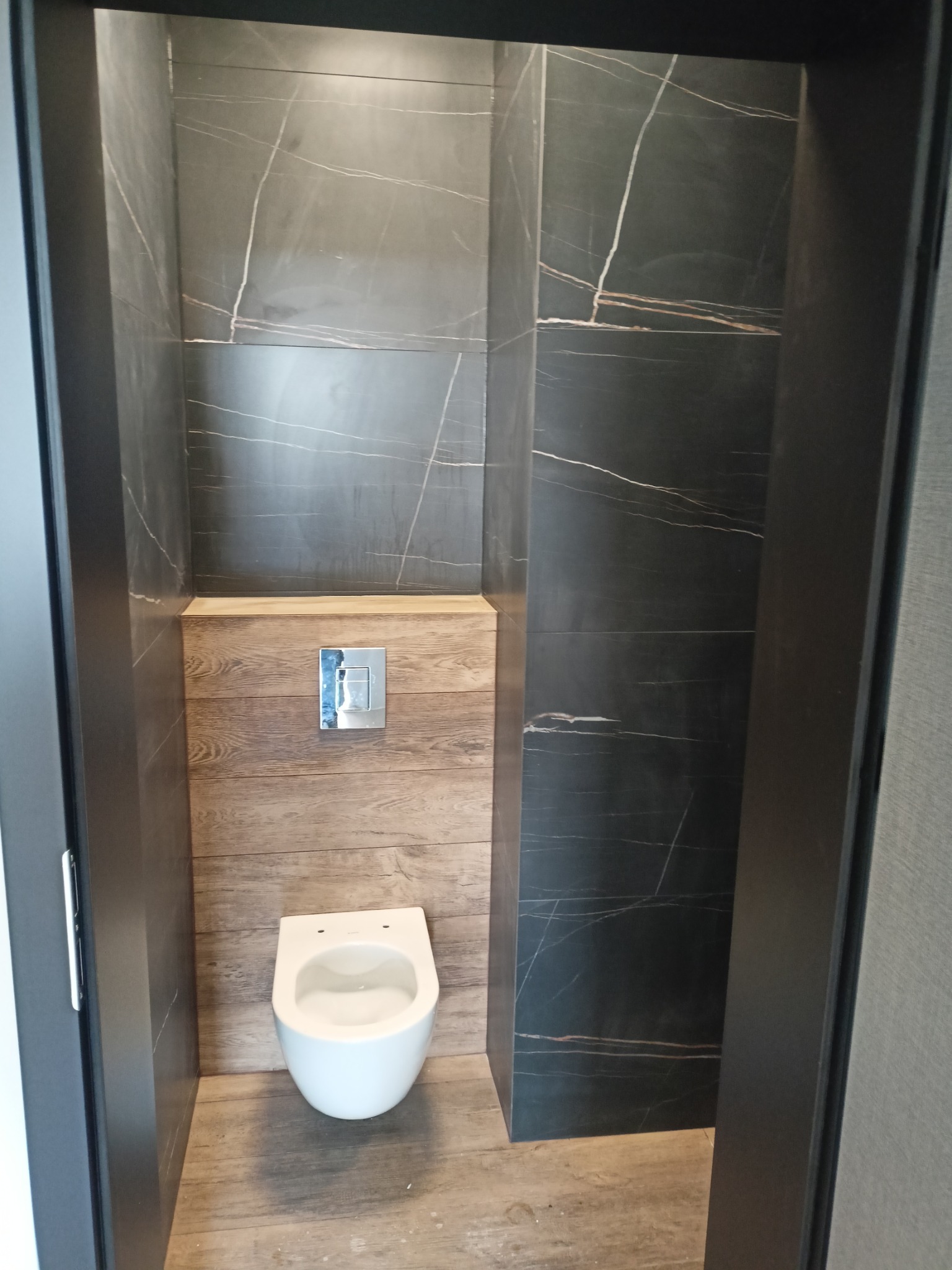 Nowoczesna toaleta w małej łazience wykończonej czarnym marmurem z białymi żyłami i drewnopodobnymi panelami, z podtynkową spłuczką.