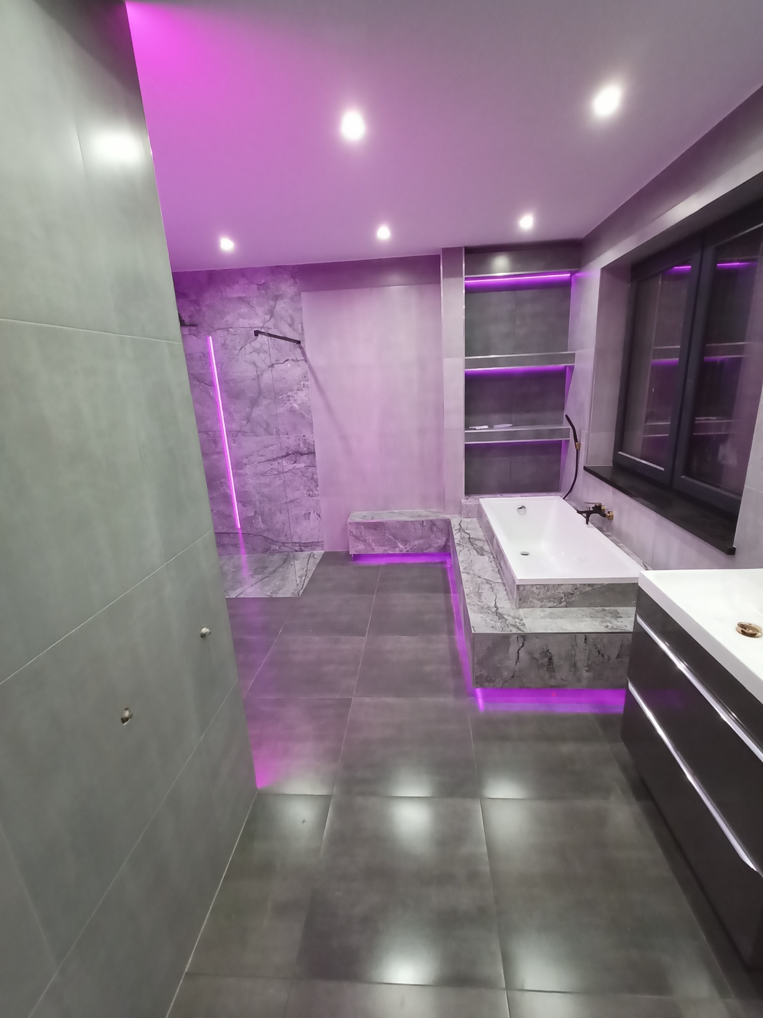 Nowoczesna łazienka z szarymi płytkami, marmurowymi akcentami i fioletowym podświetleniem LED, obejmująca kabinę prysznicową walk-in, wannę obudowaną marmurem i szafkę z umywalką.