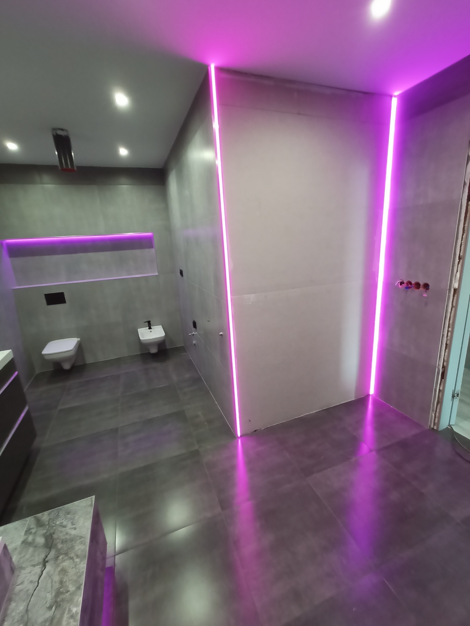Nowoczesna łazienka w szarościach z podświetleniem LED w kolorze fioletowym; widoczne płytki na podłodze i ścianach, toaleta, bidet oraz wystające instalacje wodne.