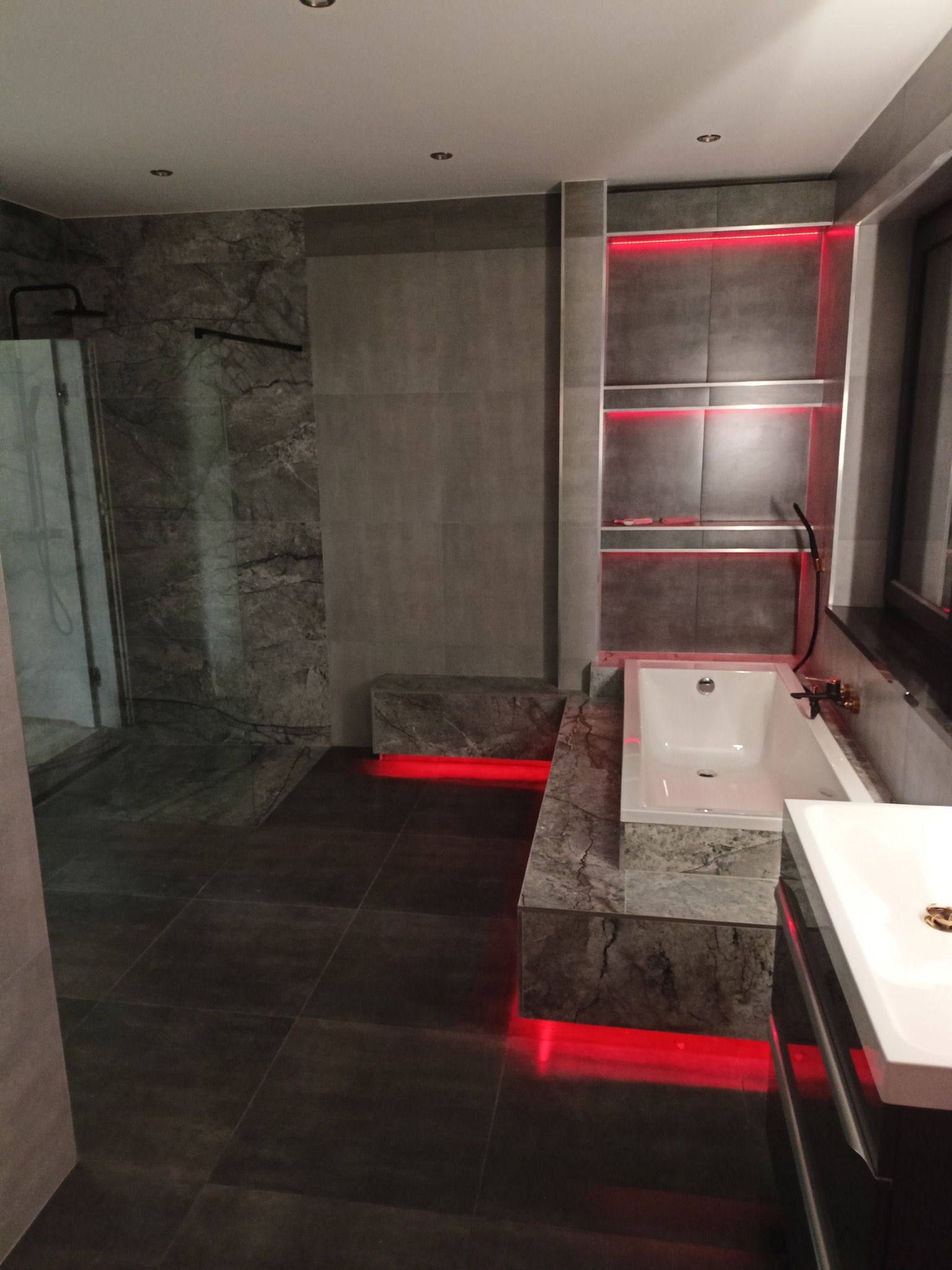 Nowoczesna łazienka z szarymi płytkami imitującymi marmur, podświetleniem LED w kolorze czerwonym pod wanną i półkami, kabiną prysznicową typu walk-in i umywalką na szafce.