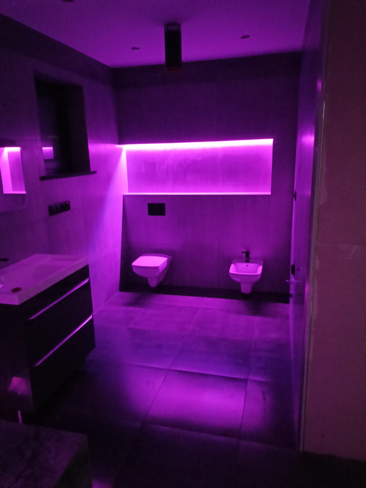 Nowoczesna łazienka z podświetleniem LED w kolorze fioletowym, widoczne umywalka z szafką, toaleta i bidet na tle szarych płytek.