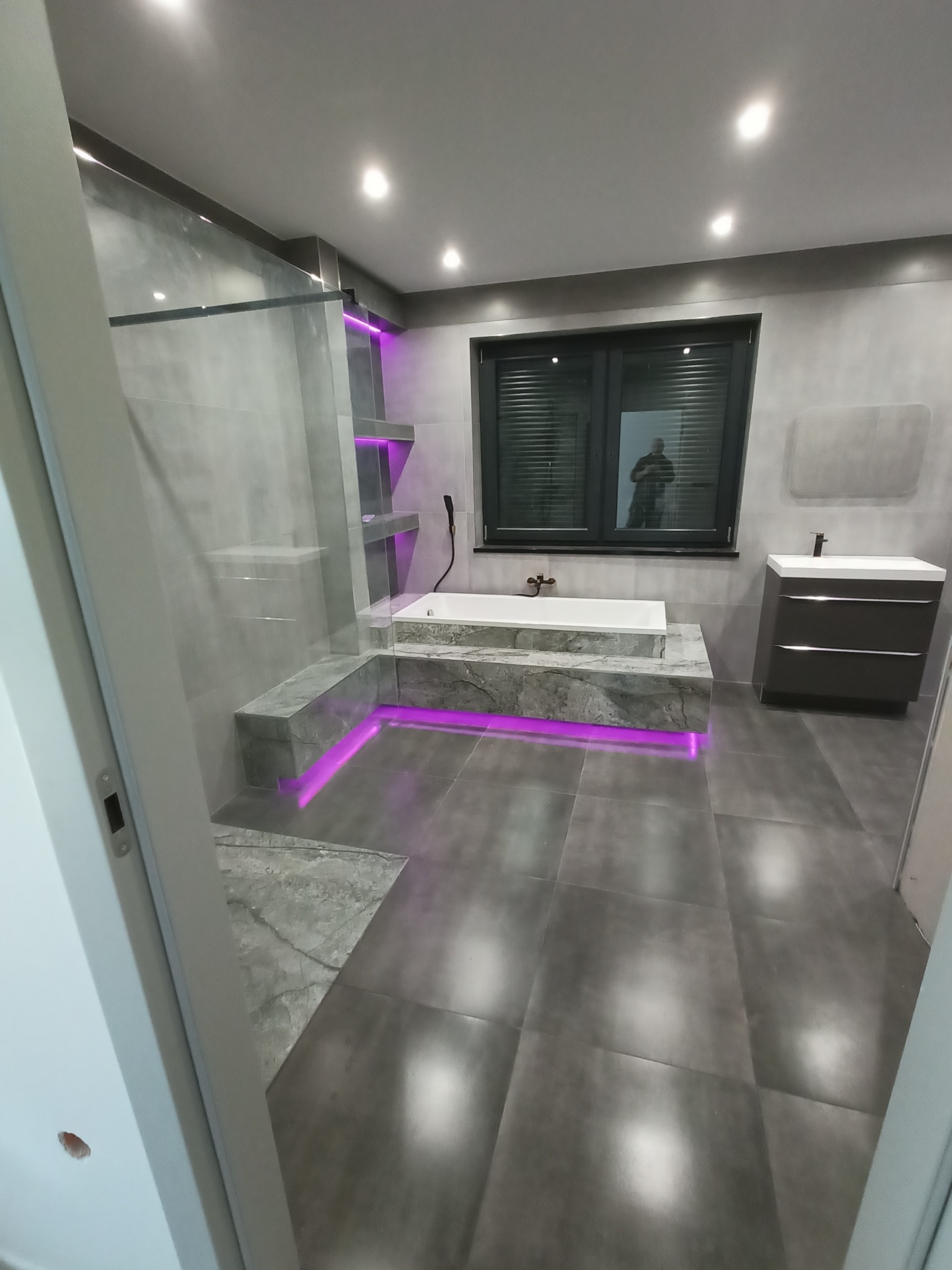 Nowoczesna łazienka z szarymi płytkami, podświetlaną wanną i kabiną prysznicową ze szklaną ścianką, widok zza otwartych drzwi.