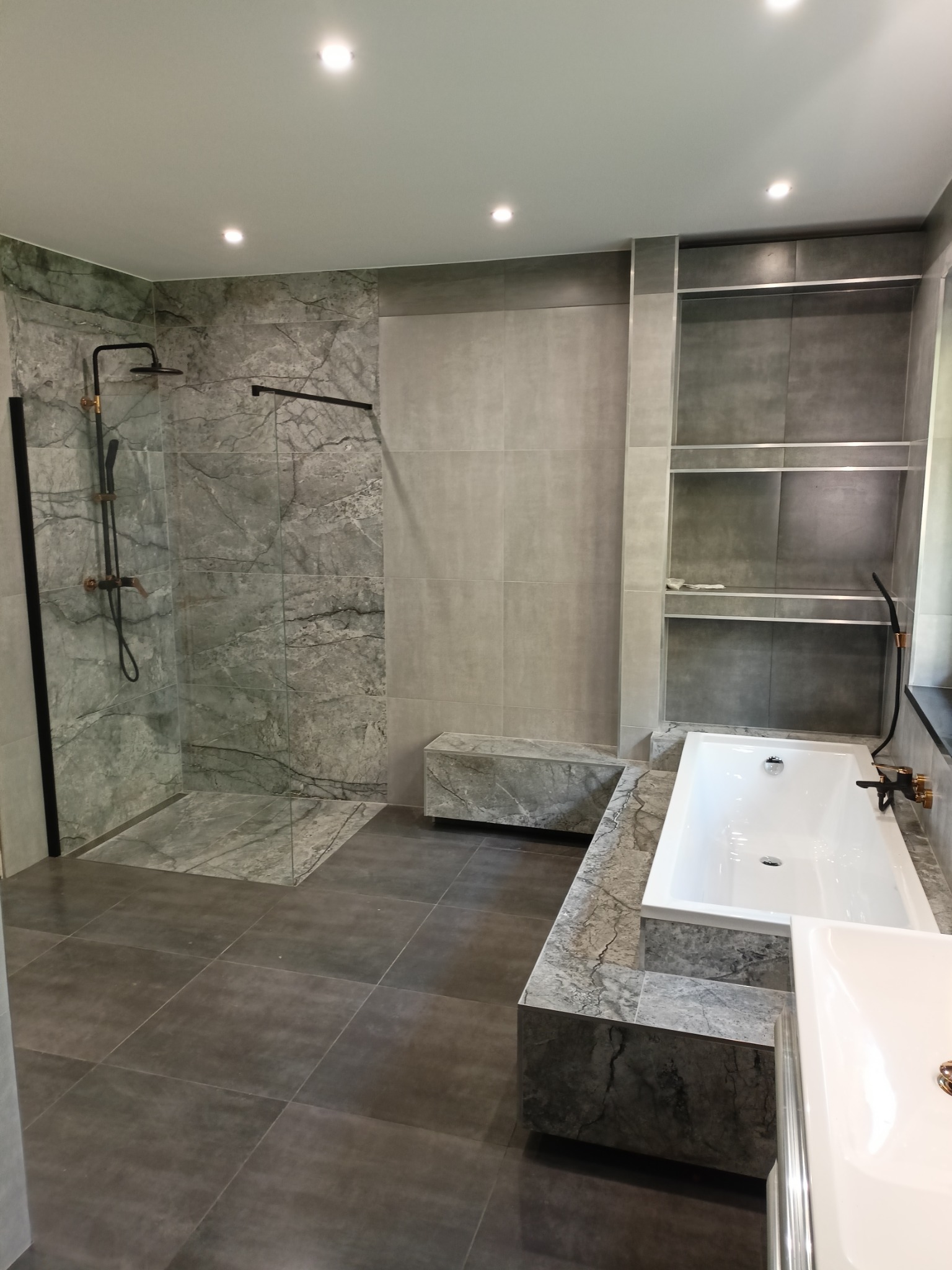 Nowoczesna łazienka z szaro-marmurkowymi płytkami, kabiną prysznicową z czarną armaturą i wanną obudowaną płytkami, oświetlona punktowymi lampami sufitowymi.