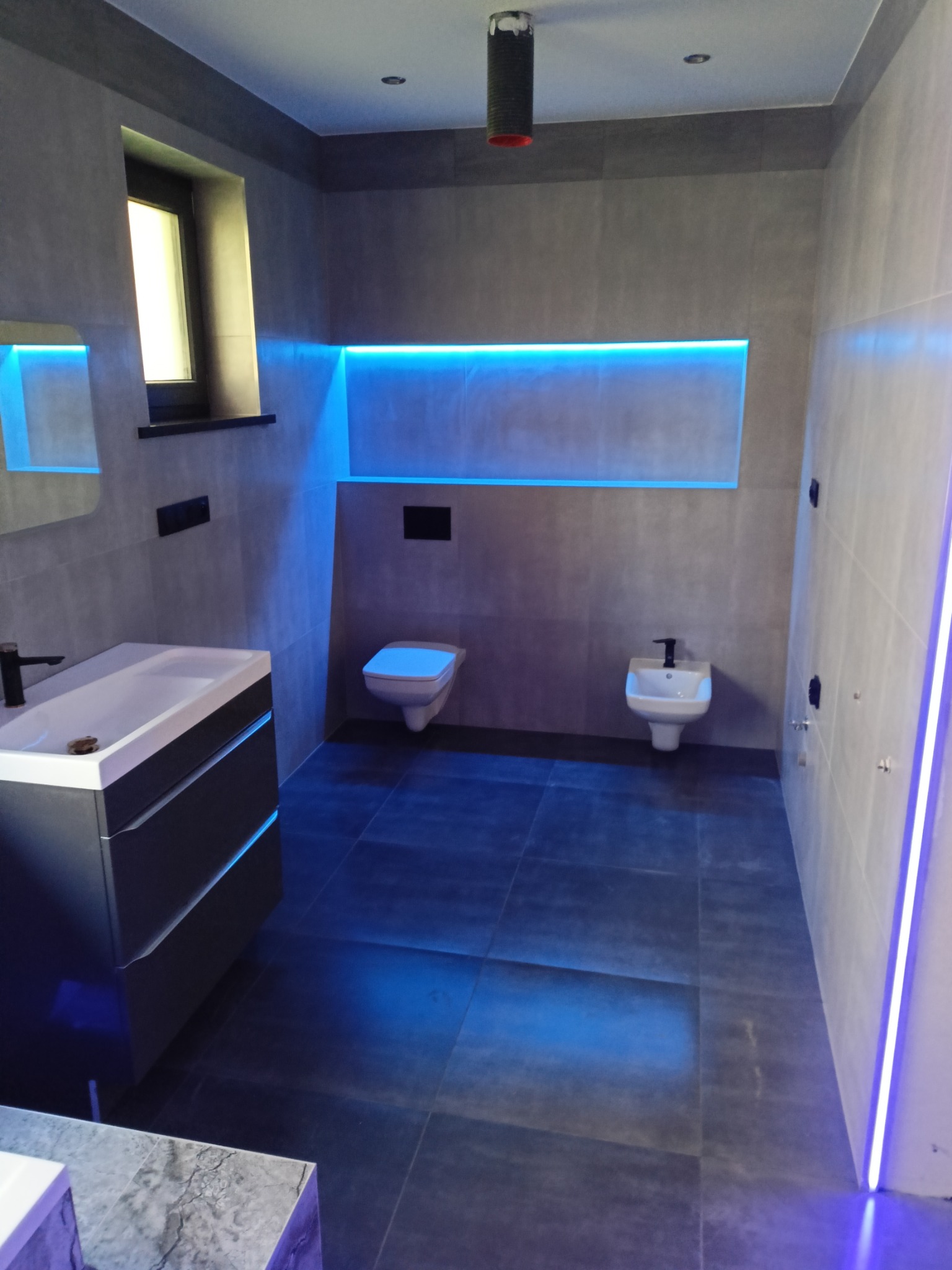 Nowoczesna łazienka z szarymi płytkami, podwieszaną toaletą i bidetem, umywalką z szafką oraz oświetleniem LED w kolorze niebieskim.