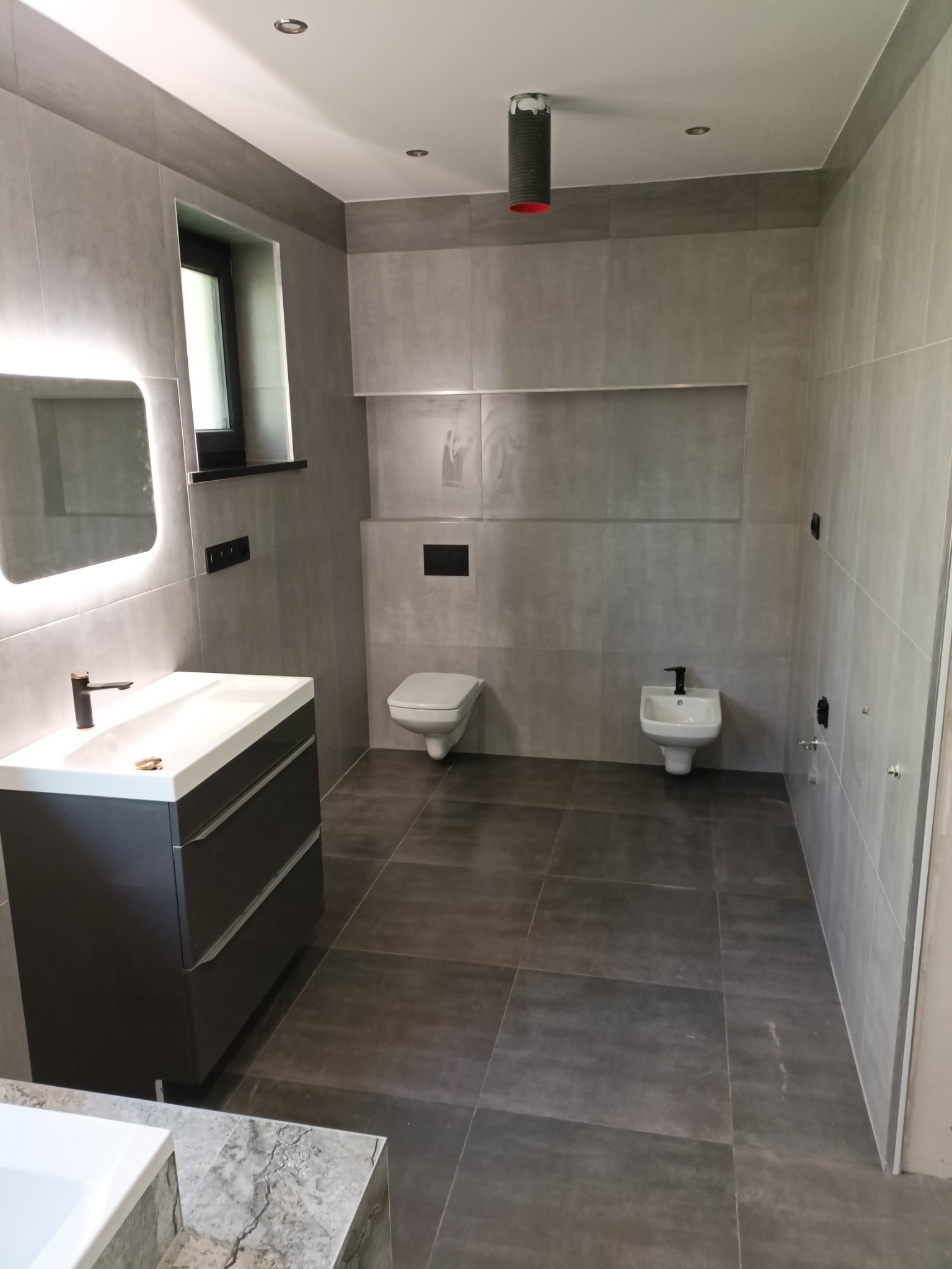 Nowoczesna łazienka w odcieniach szarości z podświetlanym lustrem, umywalką na szafce z czarną armaturą, toaletą, bidetem i ciemną, matową podłogą z płytek. Widoczne niedokończone prace przy suficie.