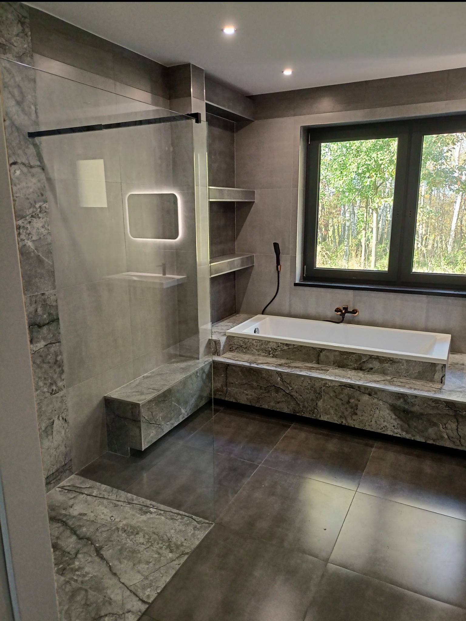 Nowoczesna łazienka z szarą marmurową okładziną, szklaną kabiną prysznicową typu walk-in, wolnostojącą wanną pod oknem i oświetlonym lustrem nad umywalką.