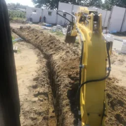 Żółta mini koparka wykonuje wąski wykop w piaszczystej glebie, w tle widoczny budynek w trakcie budowy z białych bloczków i pracujący robotnicy.