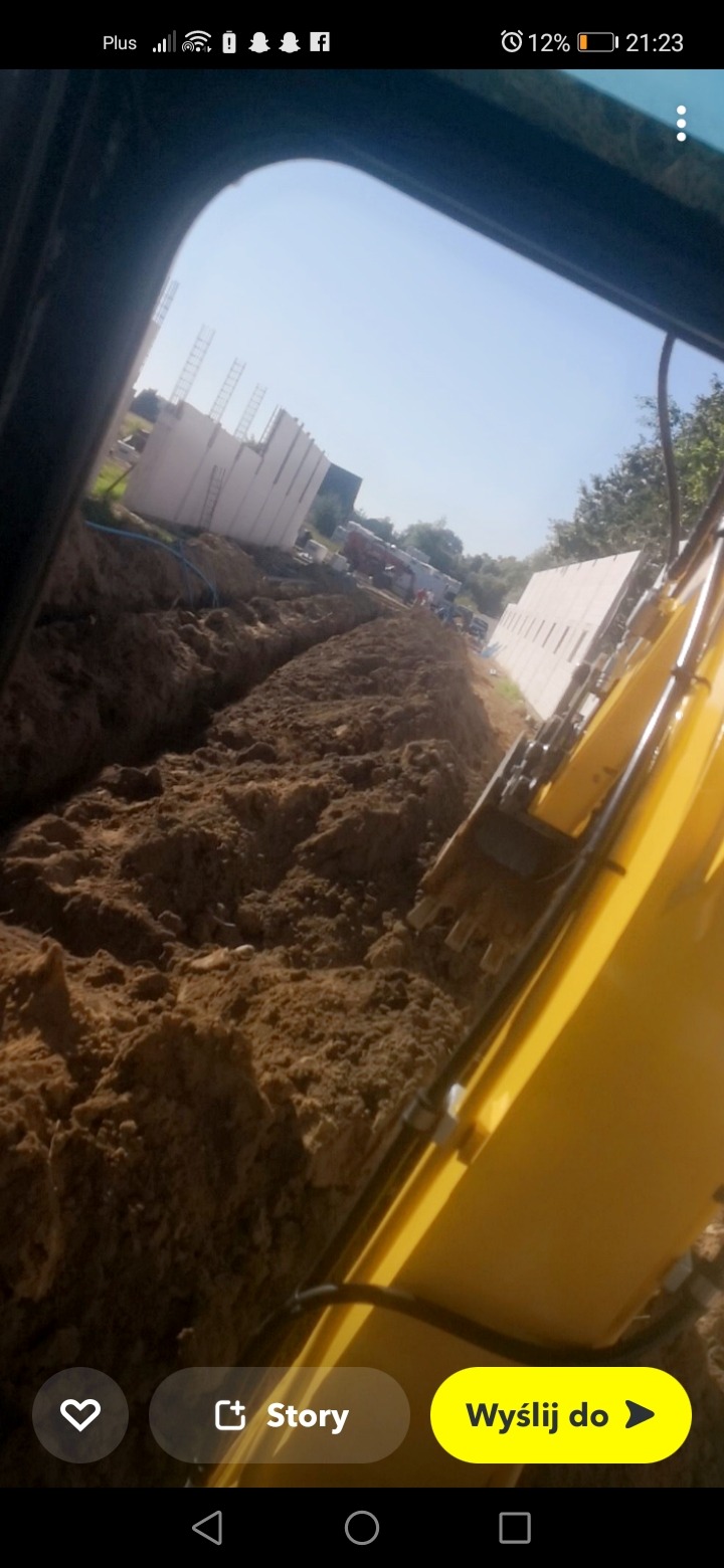Żółta koparka w trakcie wykonywania wykopu pod fundamenty, widoczne elementy szalunku i przygotowany wykop pod instalacje, zdjęcie zrobione z kabiny operatora.