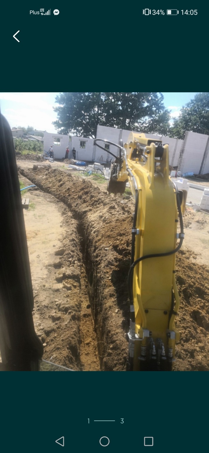 Żółta mini koparka wykonuje wąski wykop w piaszczystej glebie, w tle widoczny budynek w trakcie budowy z białych bloczków i pracujący robotnicy.