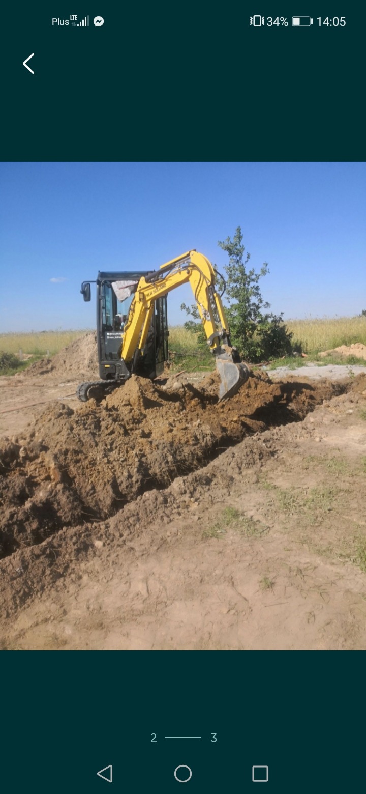 Żółto-czarna minikoparka New Holland podczas prac ziemnych, widoczny wykop w ziemi, w tle trawa i drzewa pod błękitnym niebem.