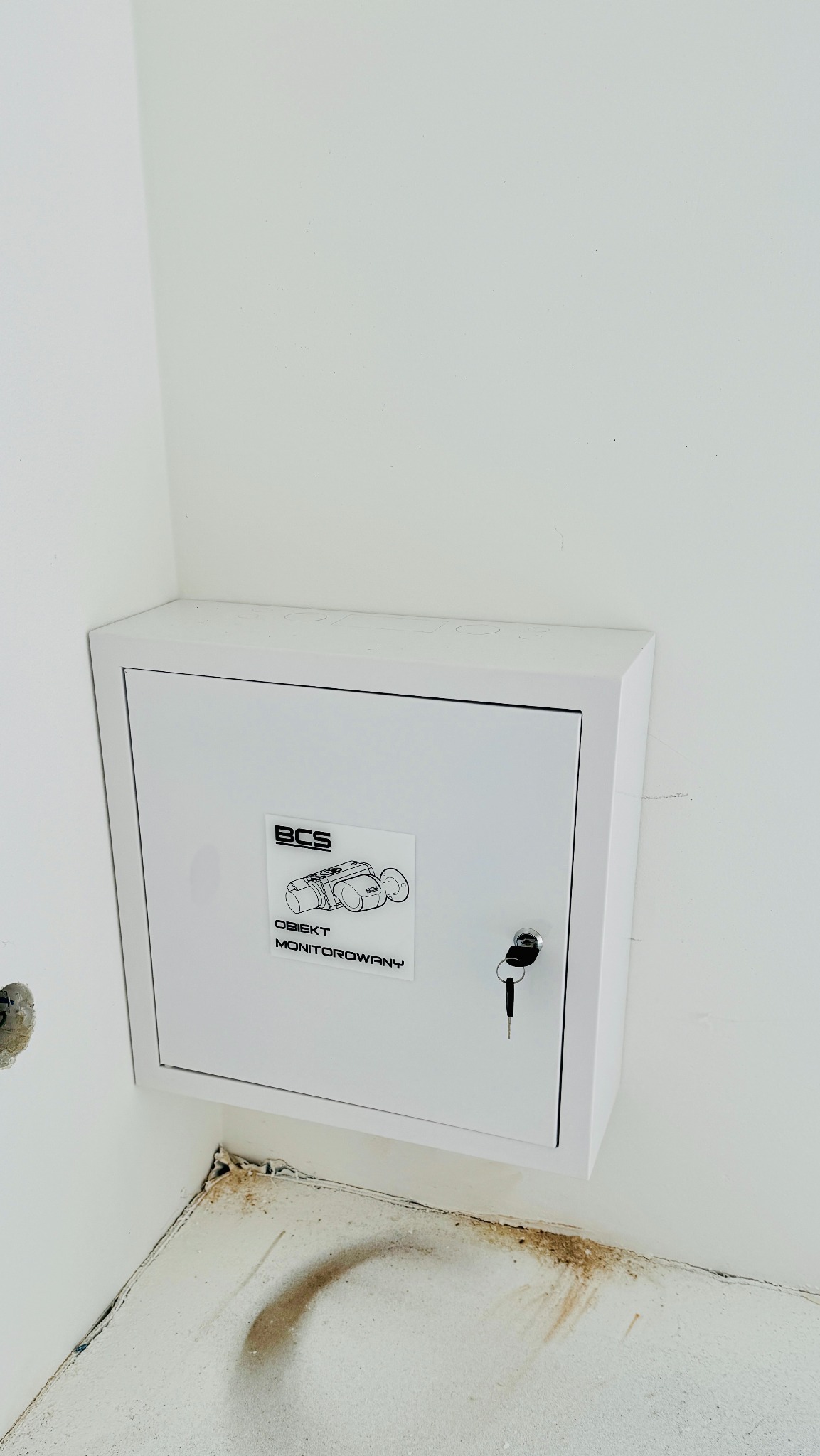 Biała szafka sterownicza systemu monitoringu BCS, zamontowana w rogu pomieszczenia w trakcie remontu, z naklejką informującą o monitoringu i kluczem w zamku.