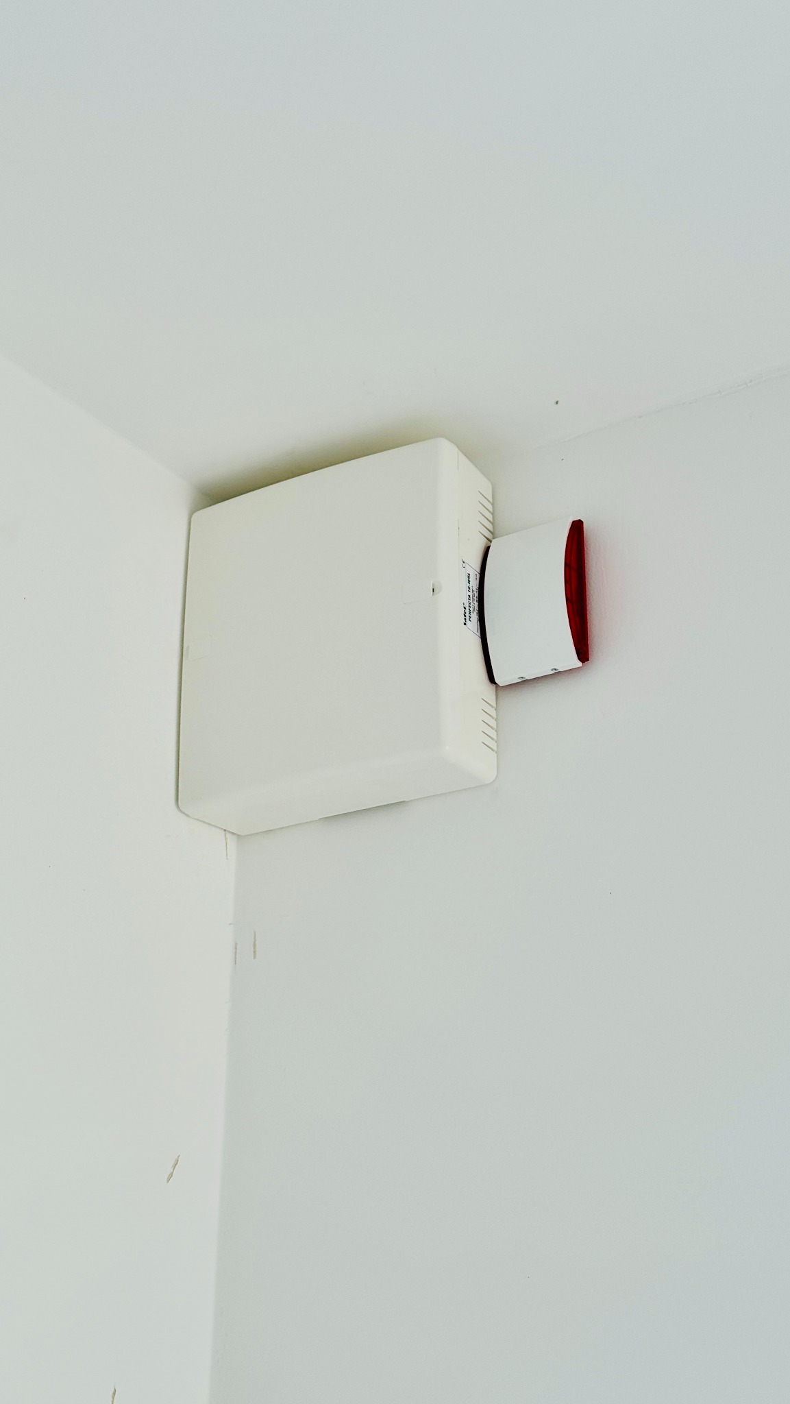 Biała centrala alarmowa z sygnalizatorem akustyczno-optycznym zamontowana w rogu pomieszczenia o białych ścianach.