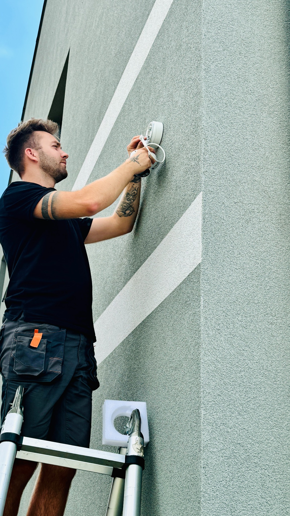 Mężczyzna na drabinie instaluje białą kamerę monitoringu na szarej fasadzie budynku z poziomymi, białymi pasami, widoczny profil twarzy, tatuaże na rękach i narzędzia w kieszeniach spodni roboczych.