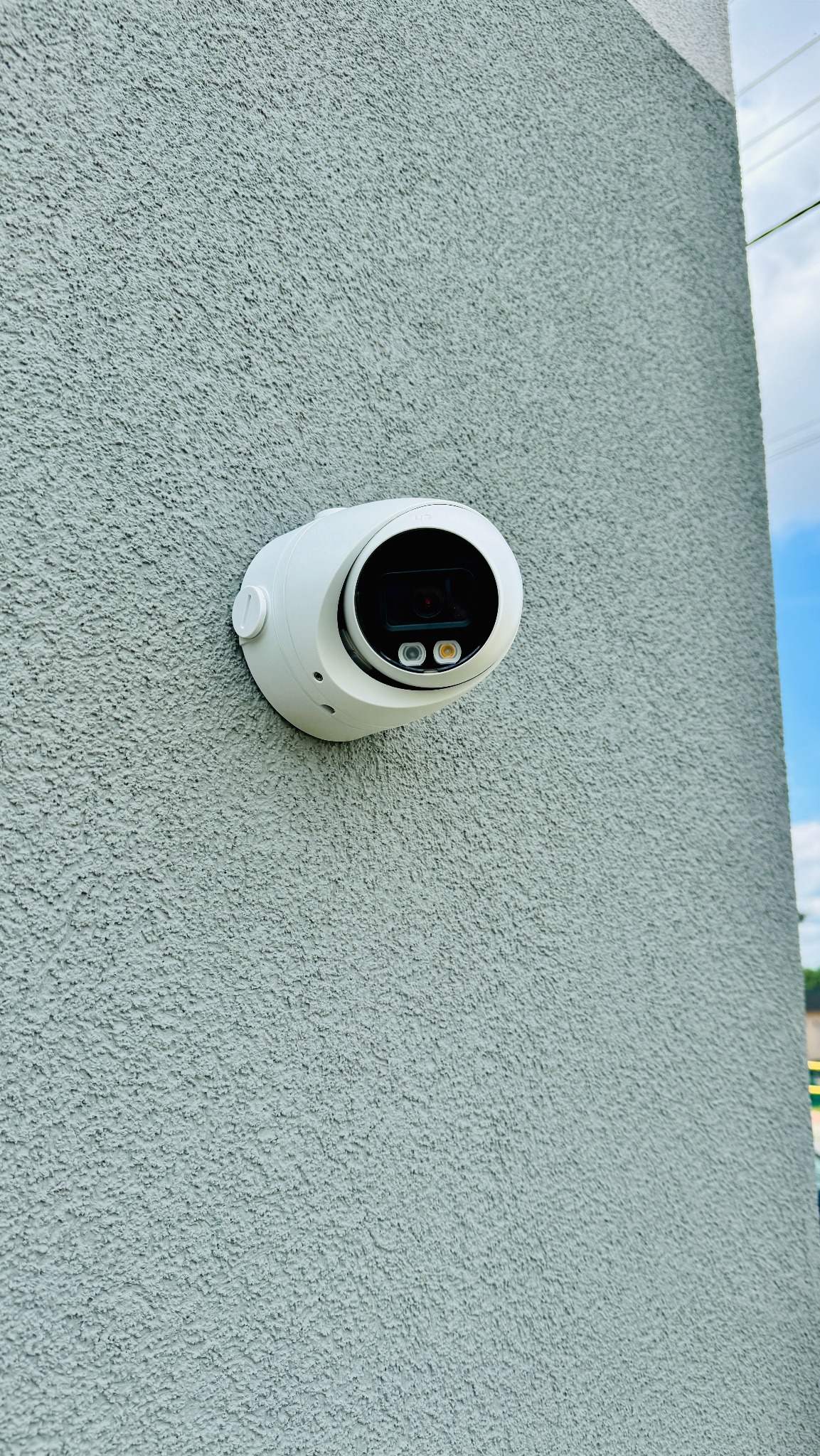 Biała kamera monitoringu zamontowana na szarej, chropowatej ścianie zewnętrznej budynku, widok z dołu.