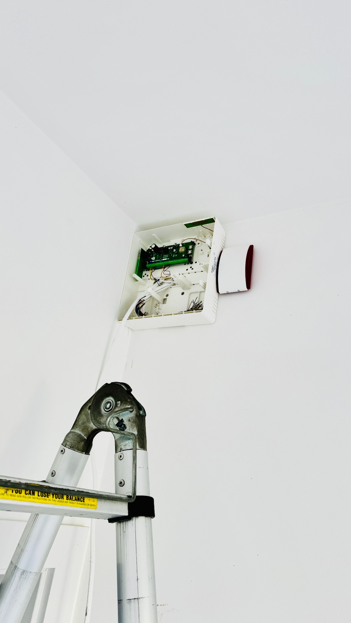 Otwarta obudowa centrali alarmowej z zieloną płytką drukowaną i podłączonymi kablami, zamontowana w rogu białego pomieszczenia, widoczna z dołu drabiny aluminiowej.