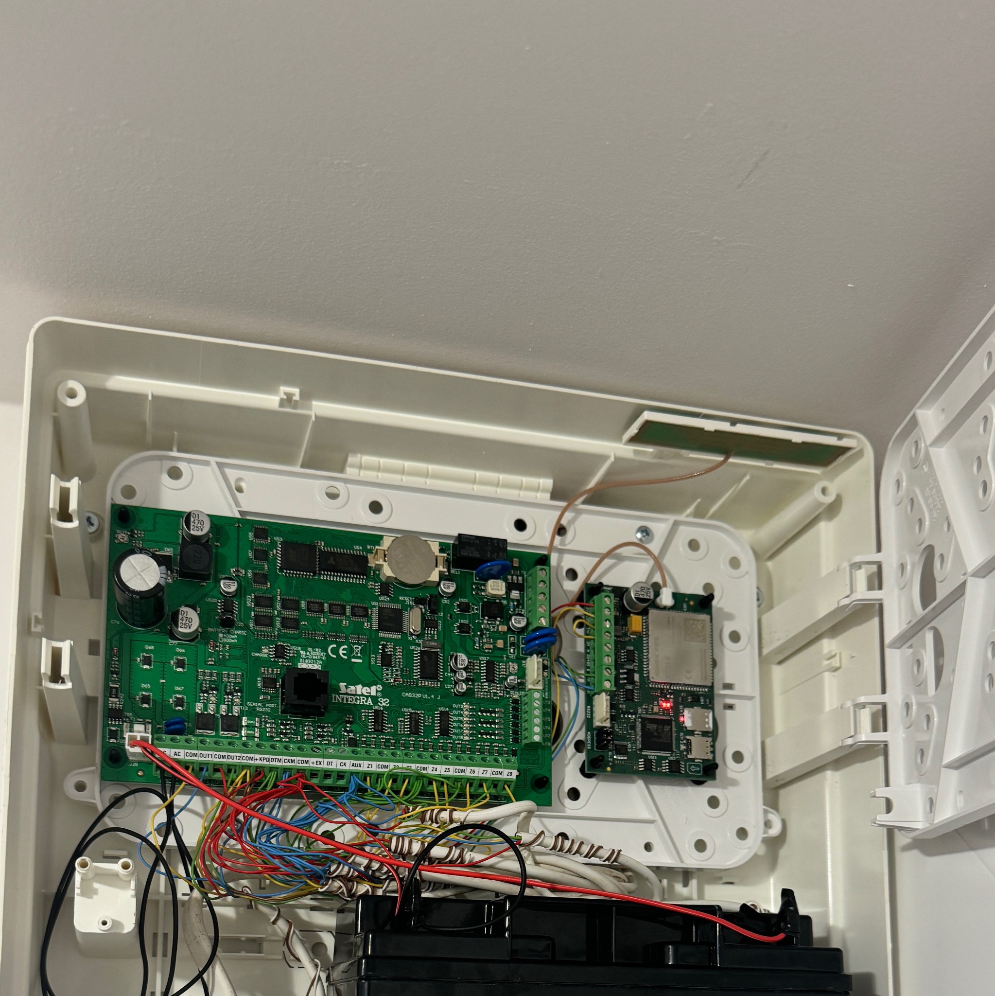 Otwarta obudowa centrali alarmowej Satel Integra 32 z widoczną płytą główną, licznymi kablami połączeniowymi w kolorach: czerwonym, niebieskim, żółtym, zielonym i białym, oraz akumulatorem awaryjnym.