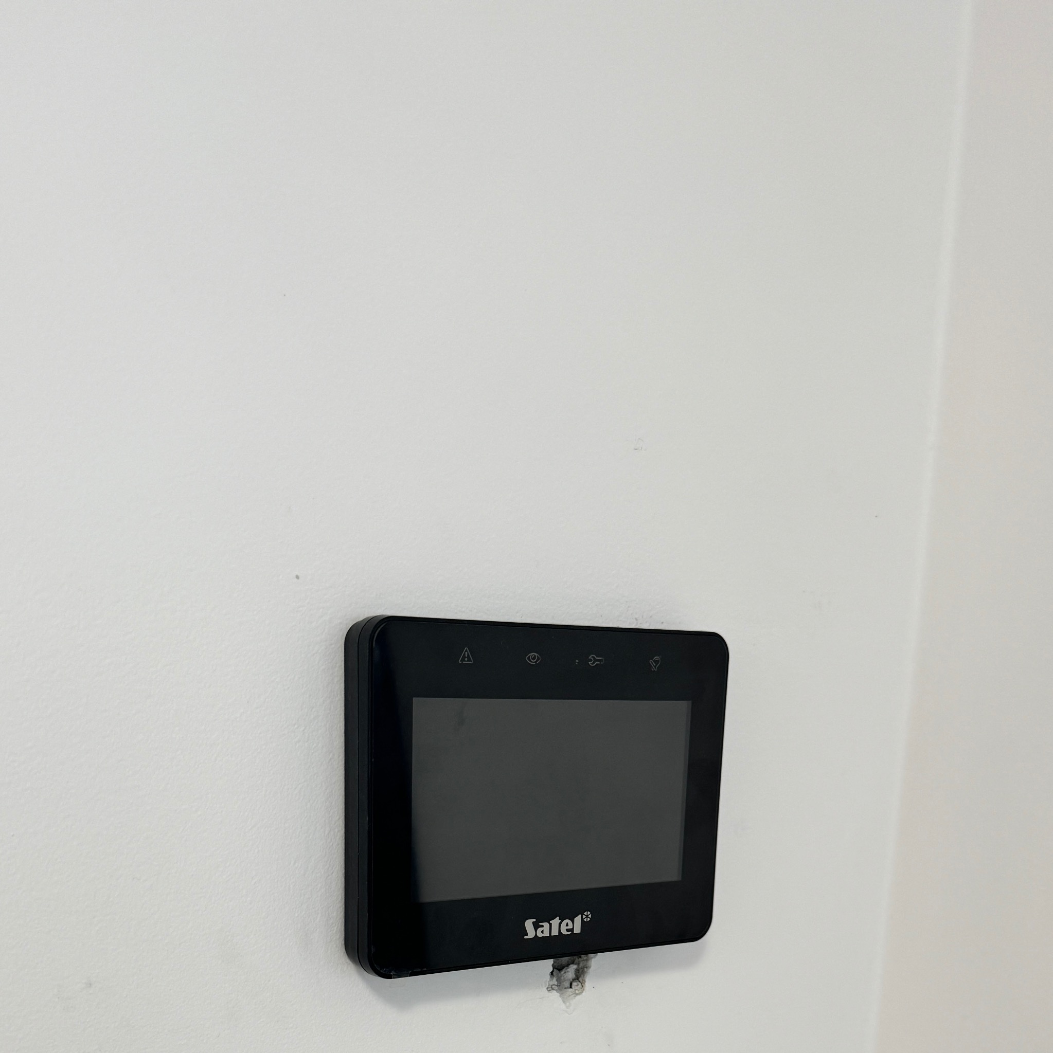 Czarny panel sterowania alarmem Satel zamontowany na białej ścianie, widoczne ślady montażu.