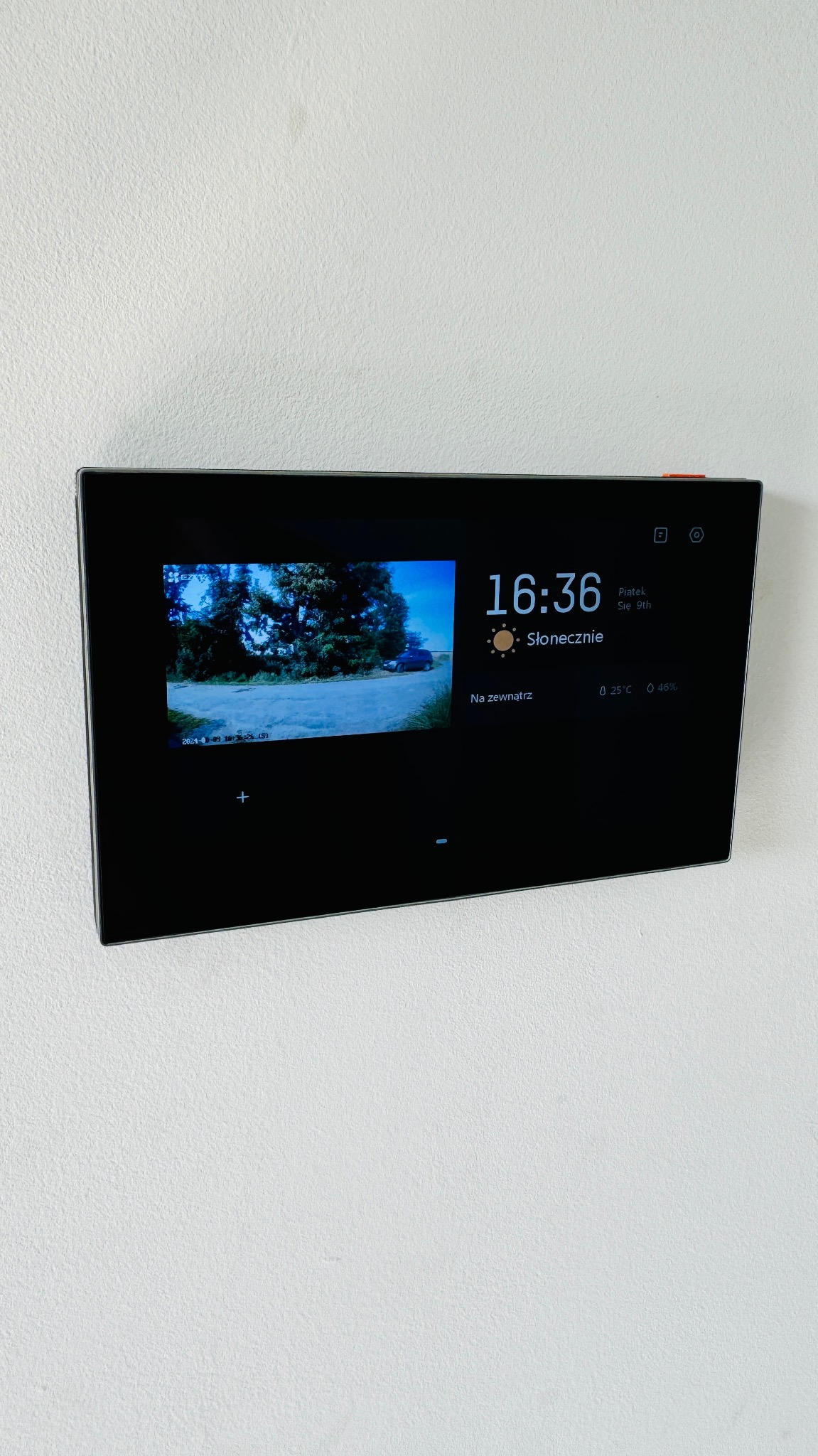 Panel dotykowy na ścianie wyświetlający obraz z kamery zewnętrznej, datę, godzinę i informacje o pogodzie.
