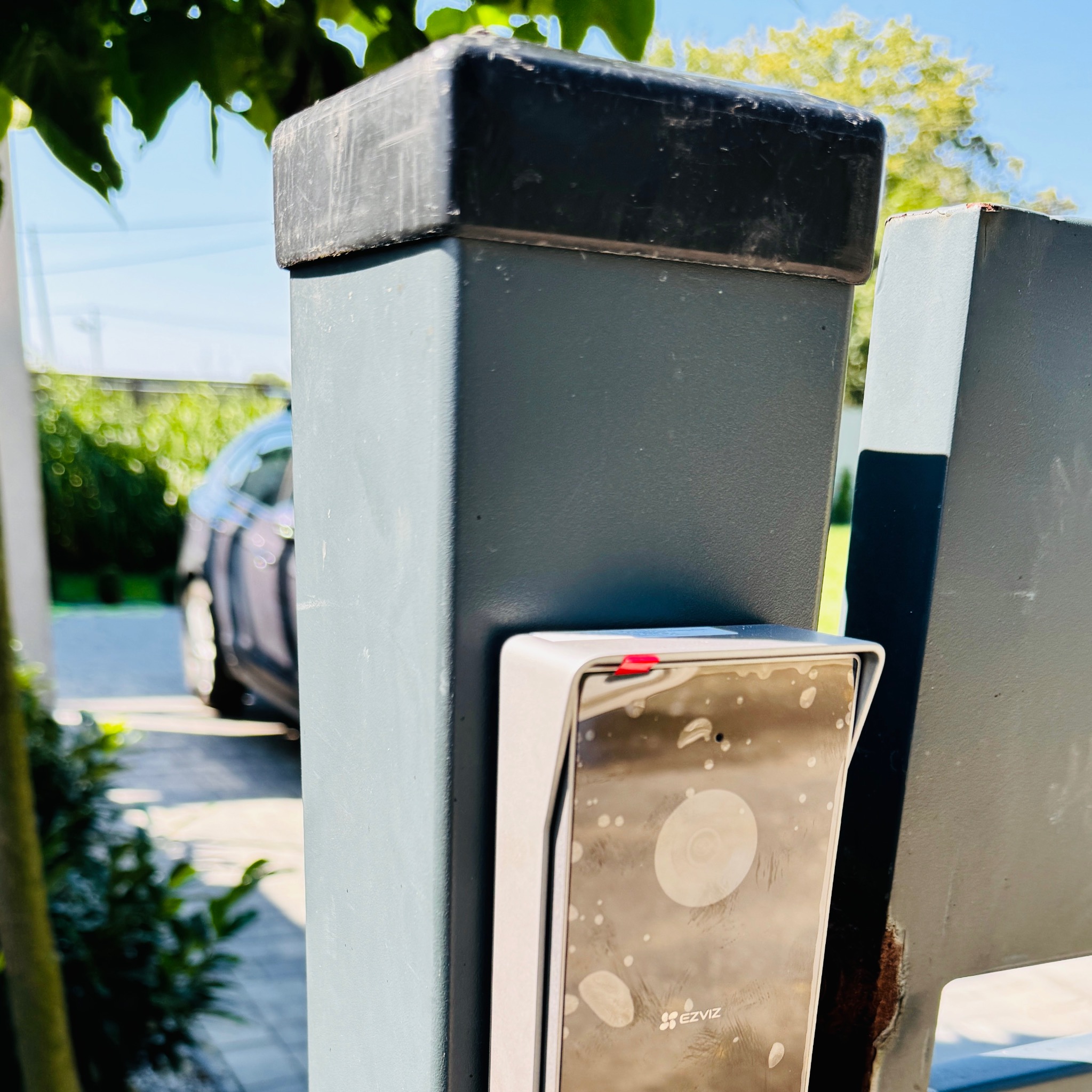 Zewnętrzny panel domofonu marki EZVIZ, zamontowany na metalowym słupku bramy wjazdowej. Widoczne ślady użytkowania i zabrudzenia na panelu. W tle rozmyty widok podjazdu i samochodu.