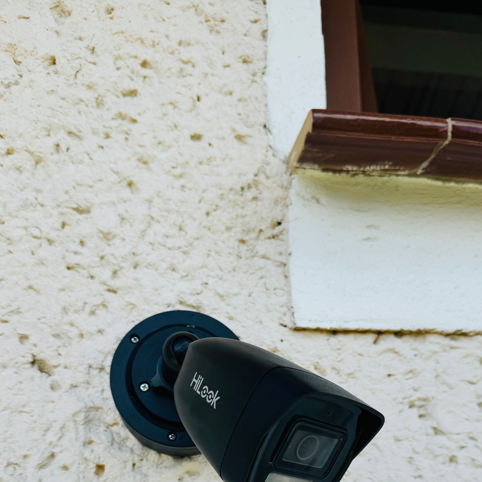 Kamera monitoringu marki HiLook zamontowana na chropowatej, jasnej ścianie budynku, widoczny fragment okna z brązową ramą.