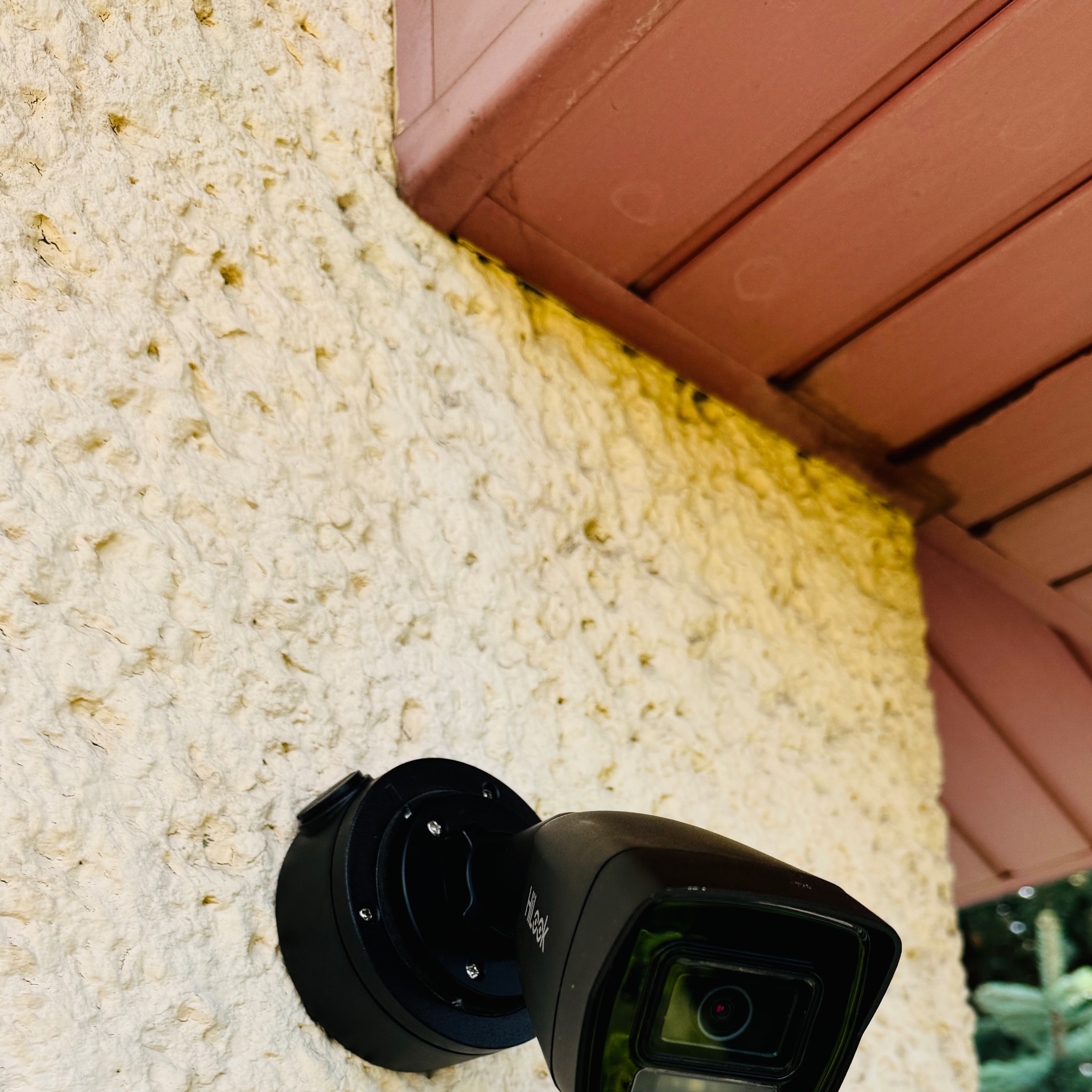 Czarna kamera monitoringu marki HiLook zamontowana na kremowej, chropowatej ścianie budynku, widoczny fragment brązowego okapu dachu.