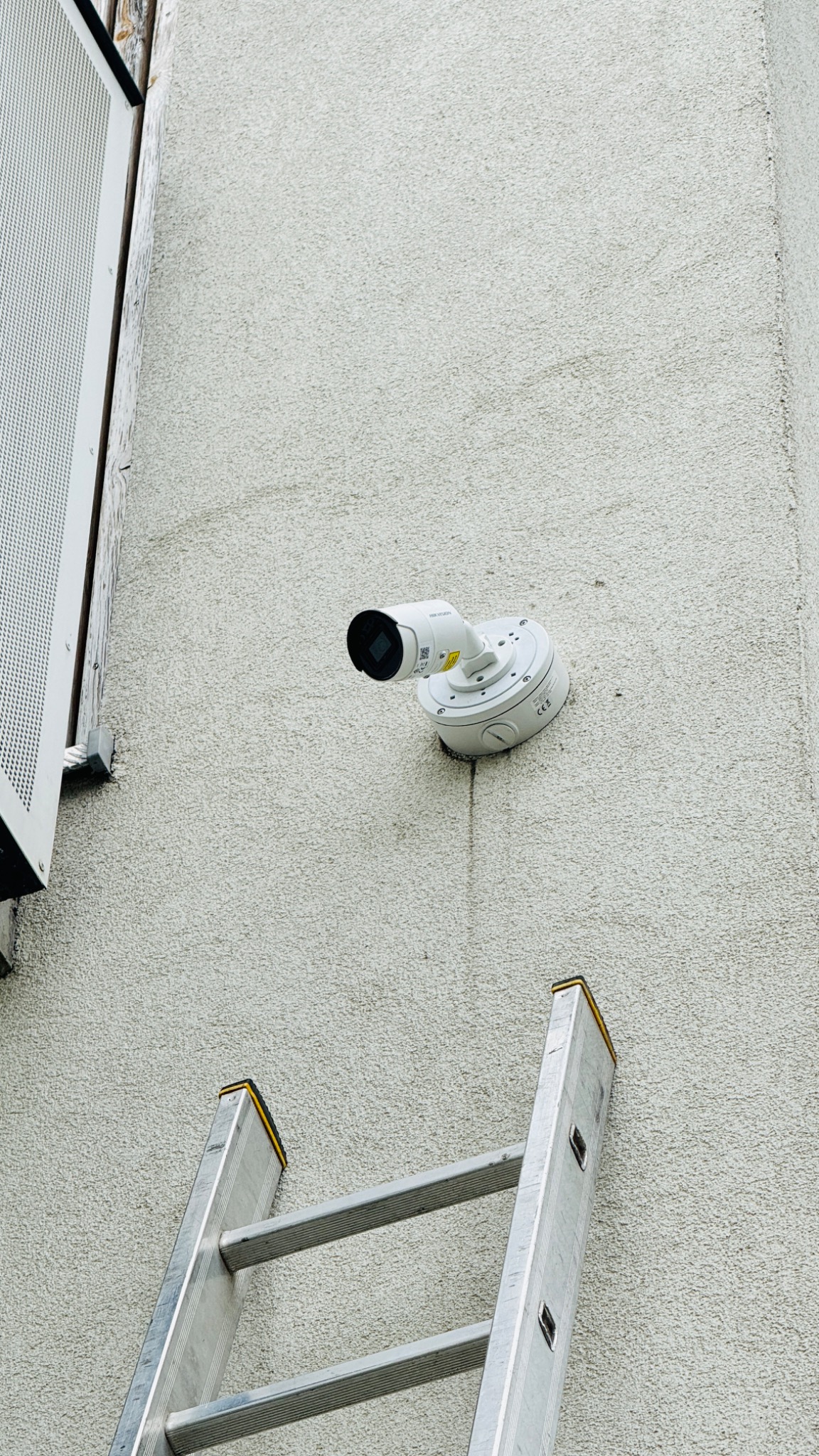 Biała kamera monitoringu Hikvision zamontowana na szarej, teksturowanej ścianie budynku, widoczna drabina aluminiowa oparta o ścianę.