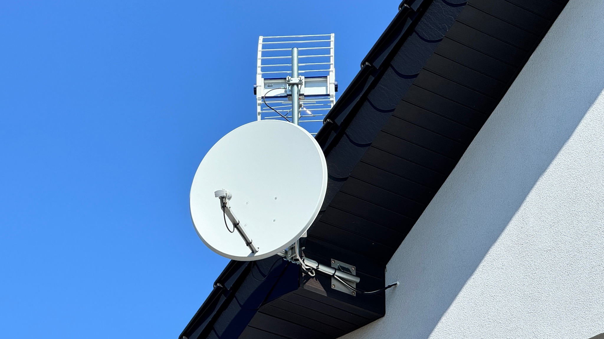 Biała antena satelitarna i antena naziemna zamontowane na czarnym dachu domu z białą elewacją na tle błękitnego nieba.