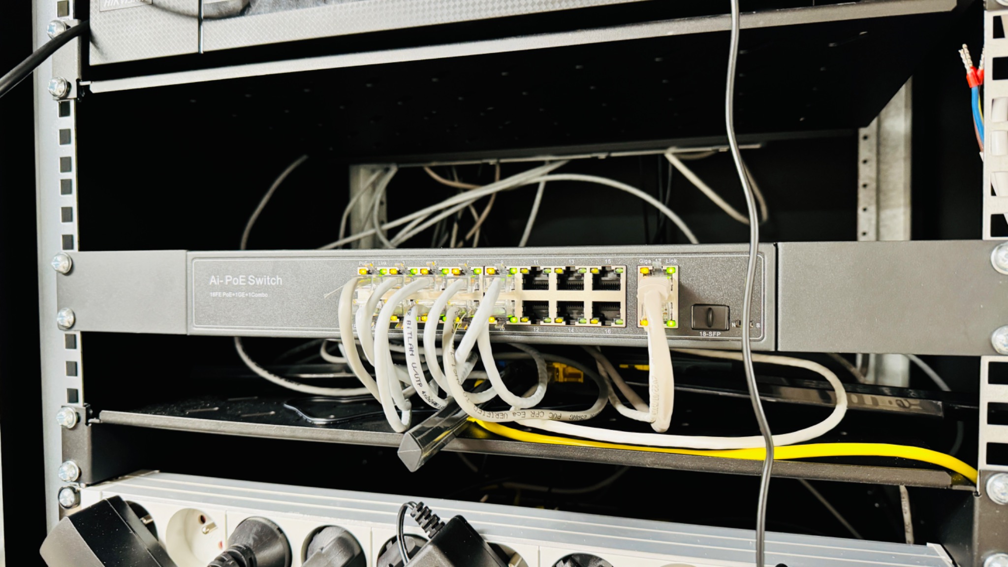 Szary przełącznik Ai-PoE Switch zamontowany w szafie rack, z podłączonymi białymi kablami Ethernet, sygnalizującymi aktywność portów.